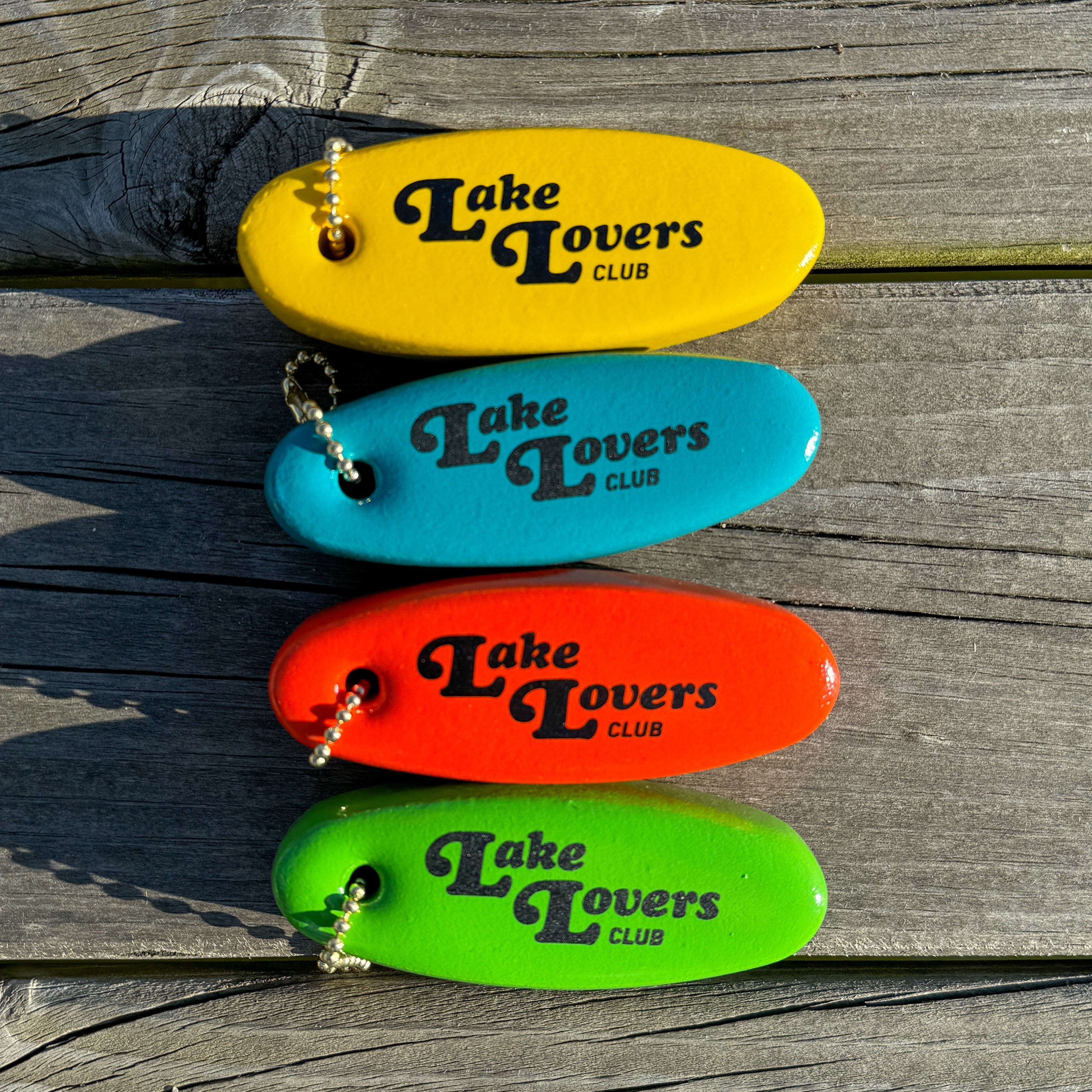 Four colorful floater key chains with 'Lake Lovers Club' branding on a wooden dock.