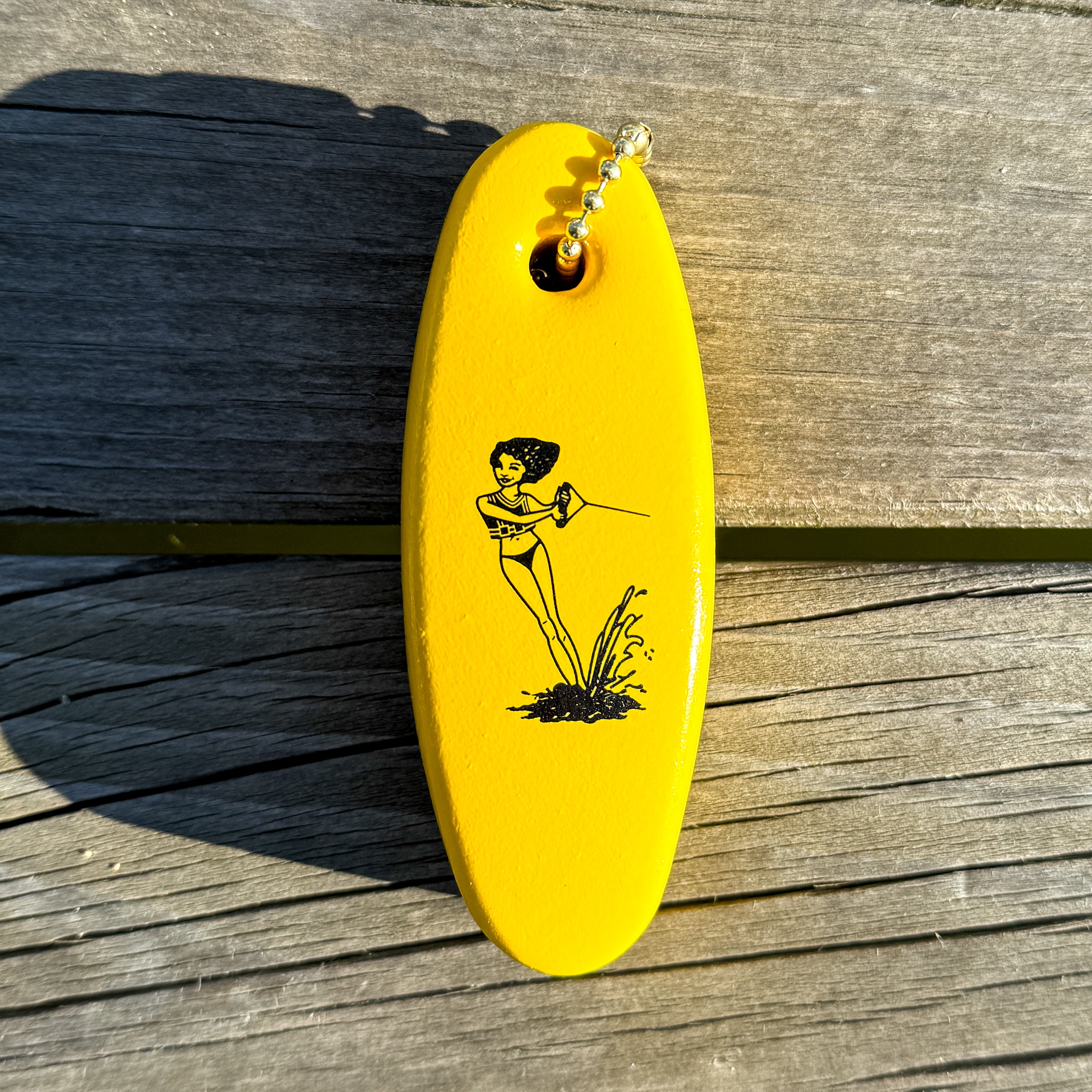 Yellow floater keychain with a black design of a girl waterskiing, lying on a wooden dock surface