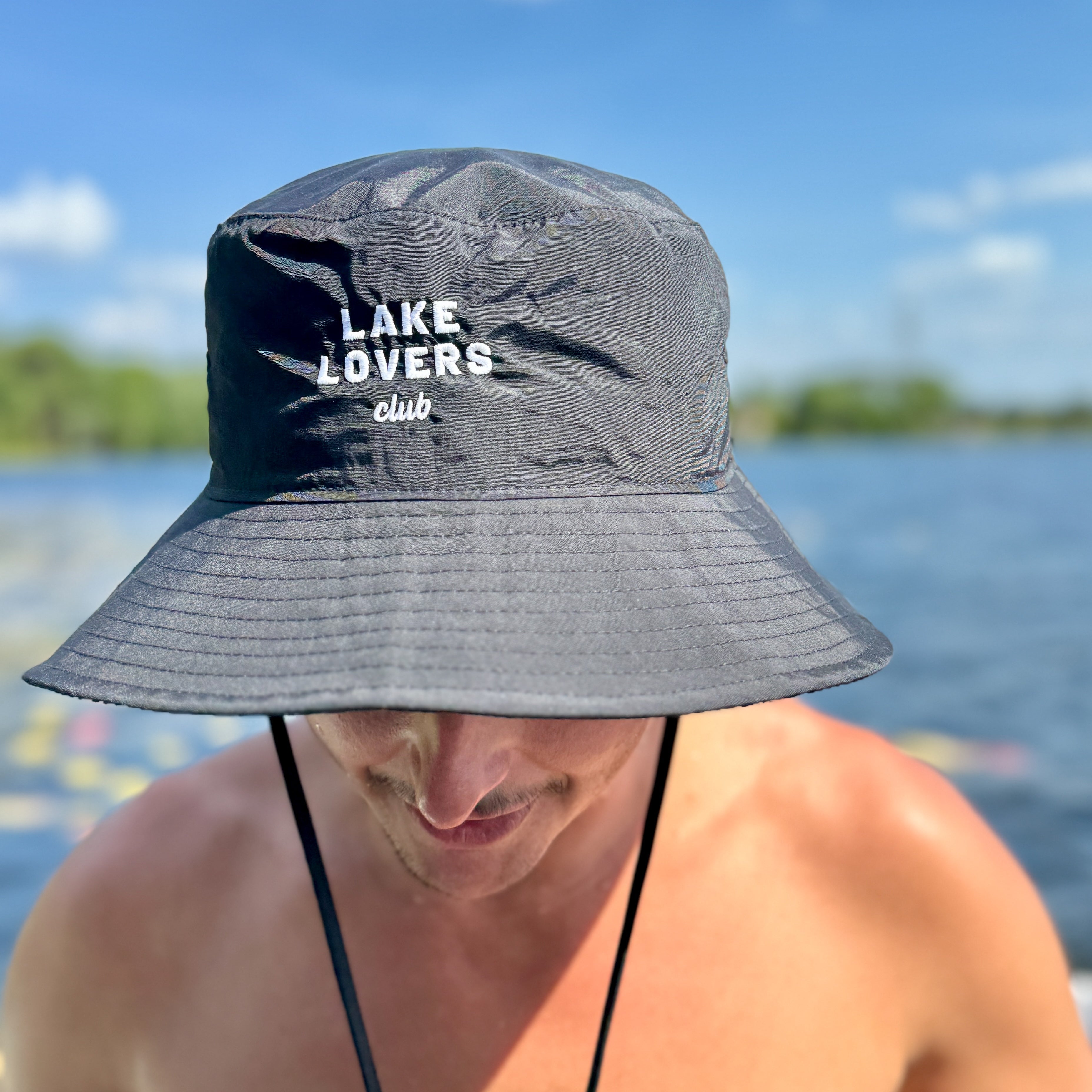 Person wearing a black bucket hat with 'Lake Lovers Club' text by a body of water.