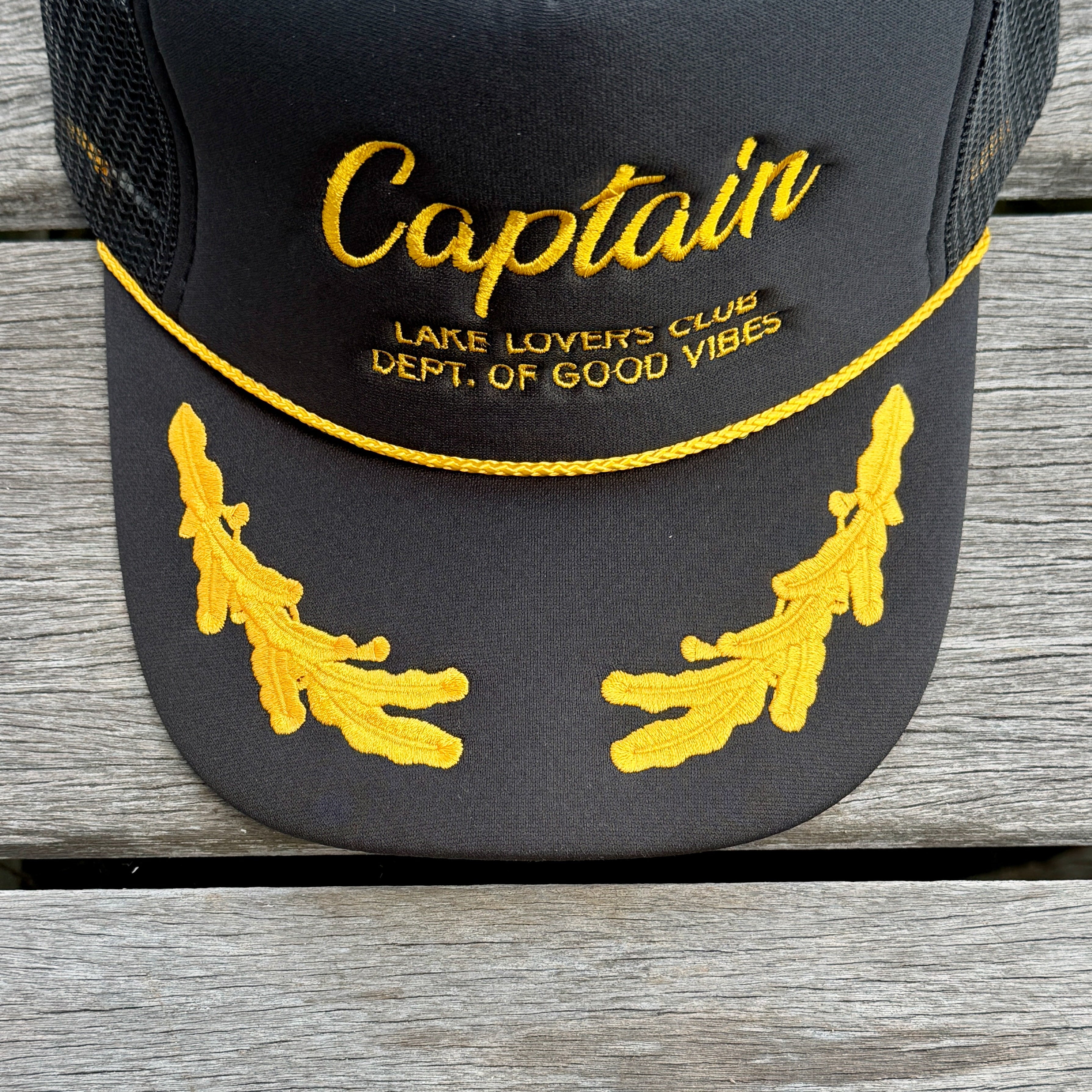 Black cap with yellow 'Captain' text and leaf designs on a wooden surface
