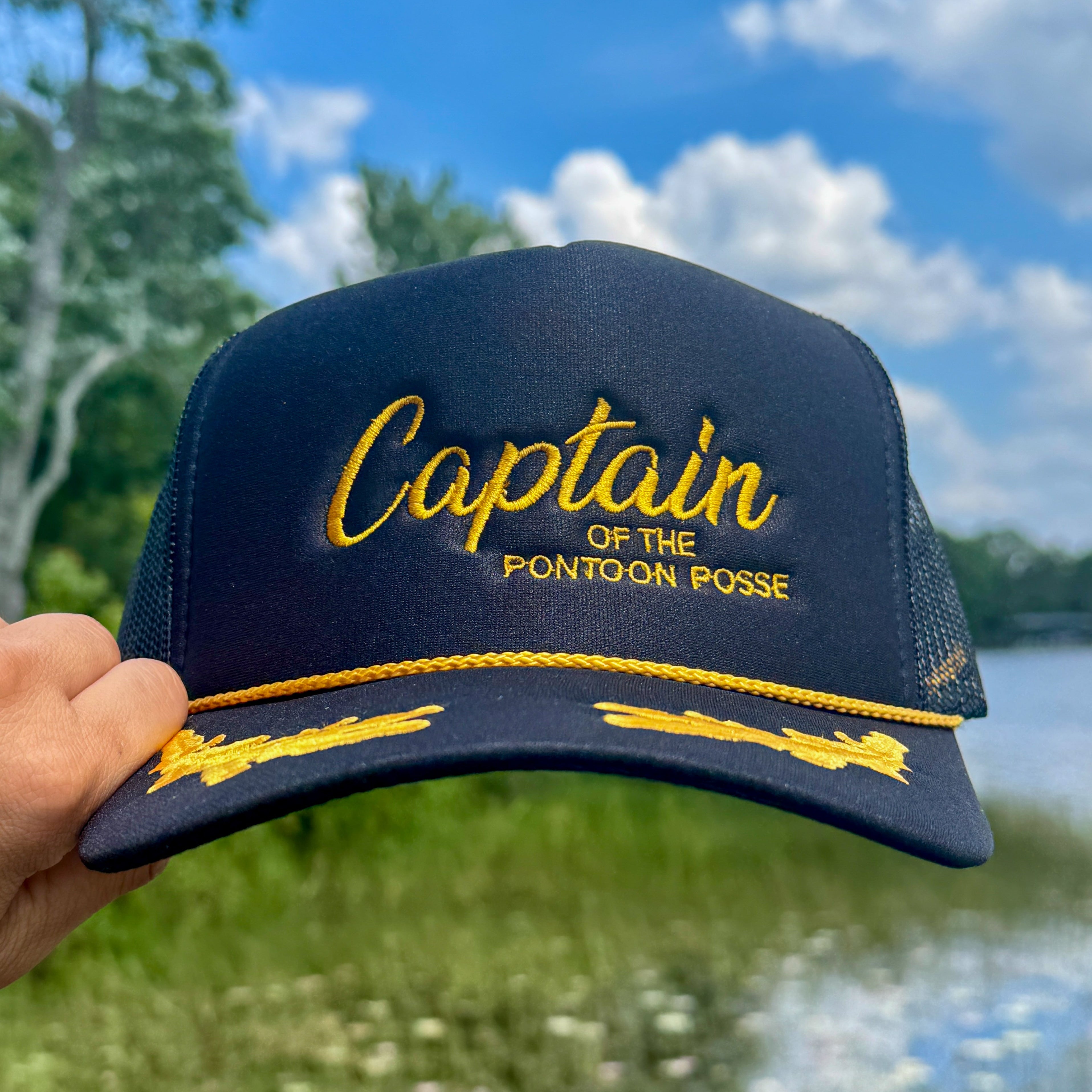 black trucker cap with yellow text 'Captain of the Pontoon Posse' held outdoors with a lake and trees in the background.