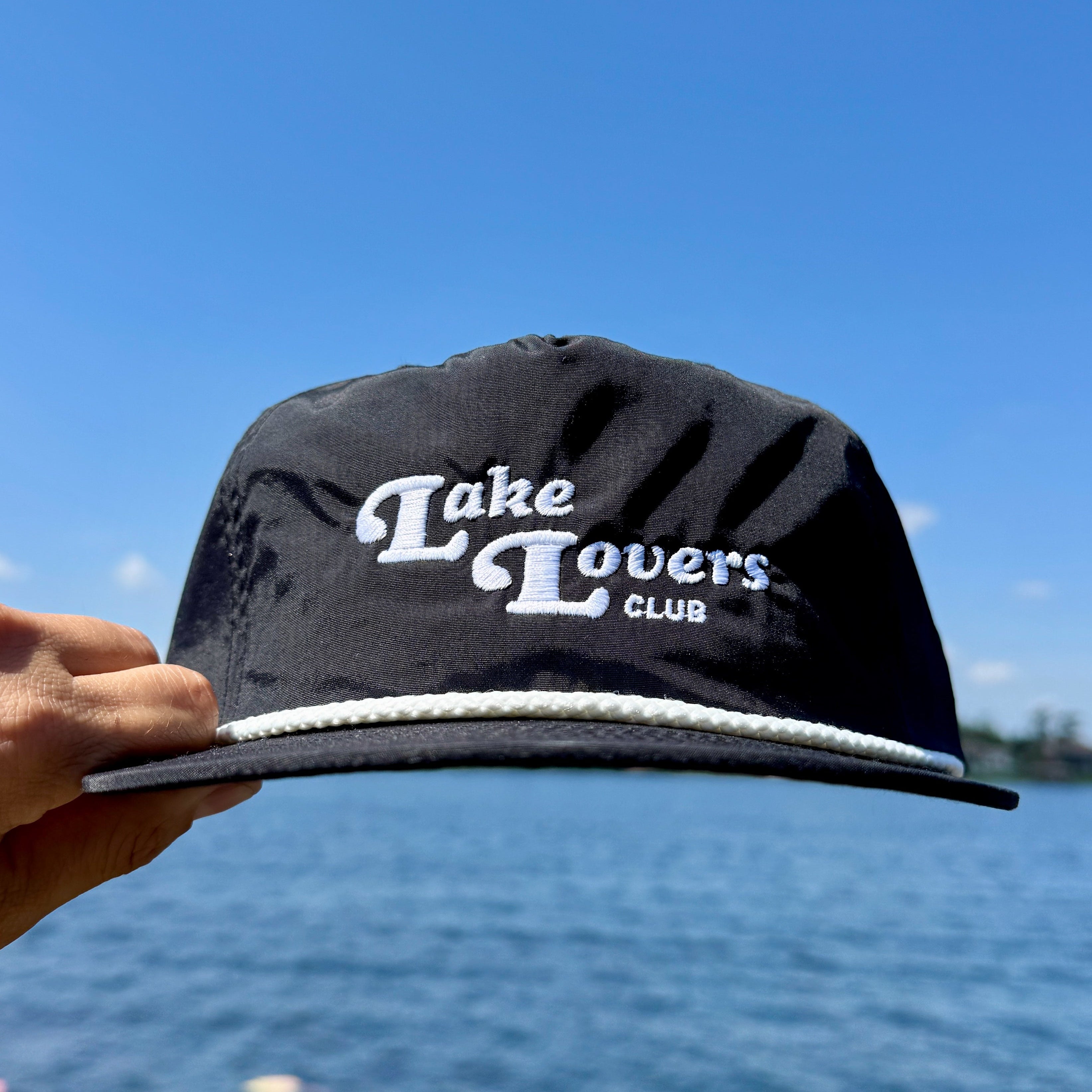 Black cap with 'Lake Lovers Club' text held against a blue sky and water background