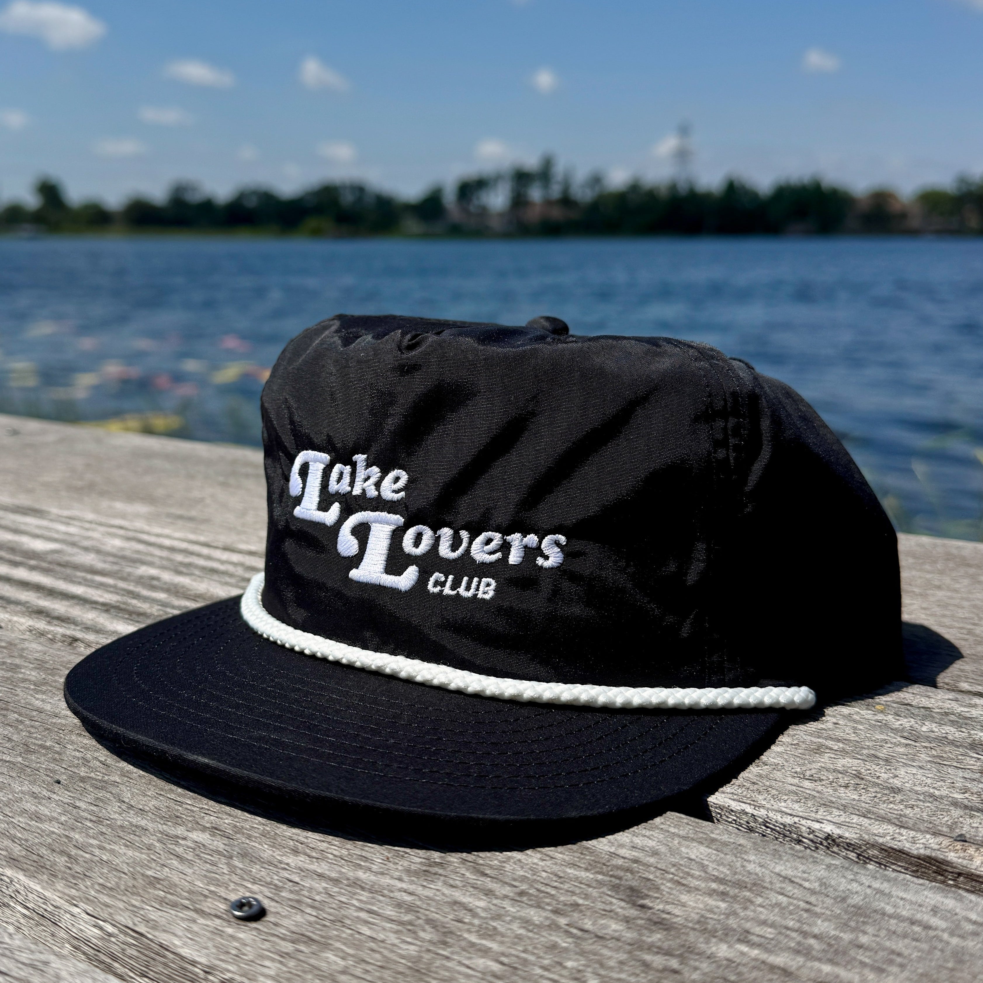 Black cap with 'Lake Lovers Club' text on a wooden surface by a lake.