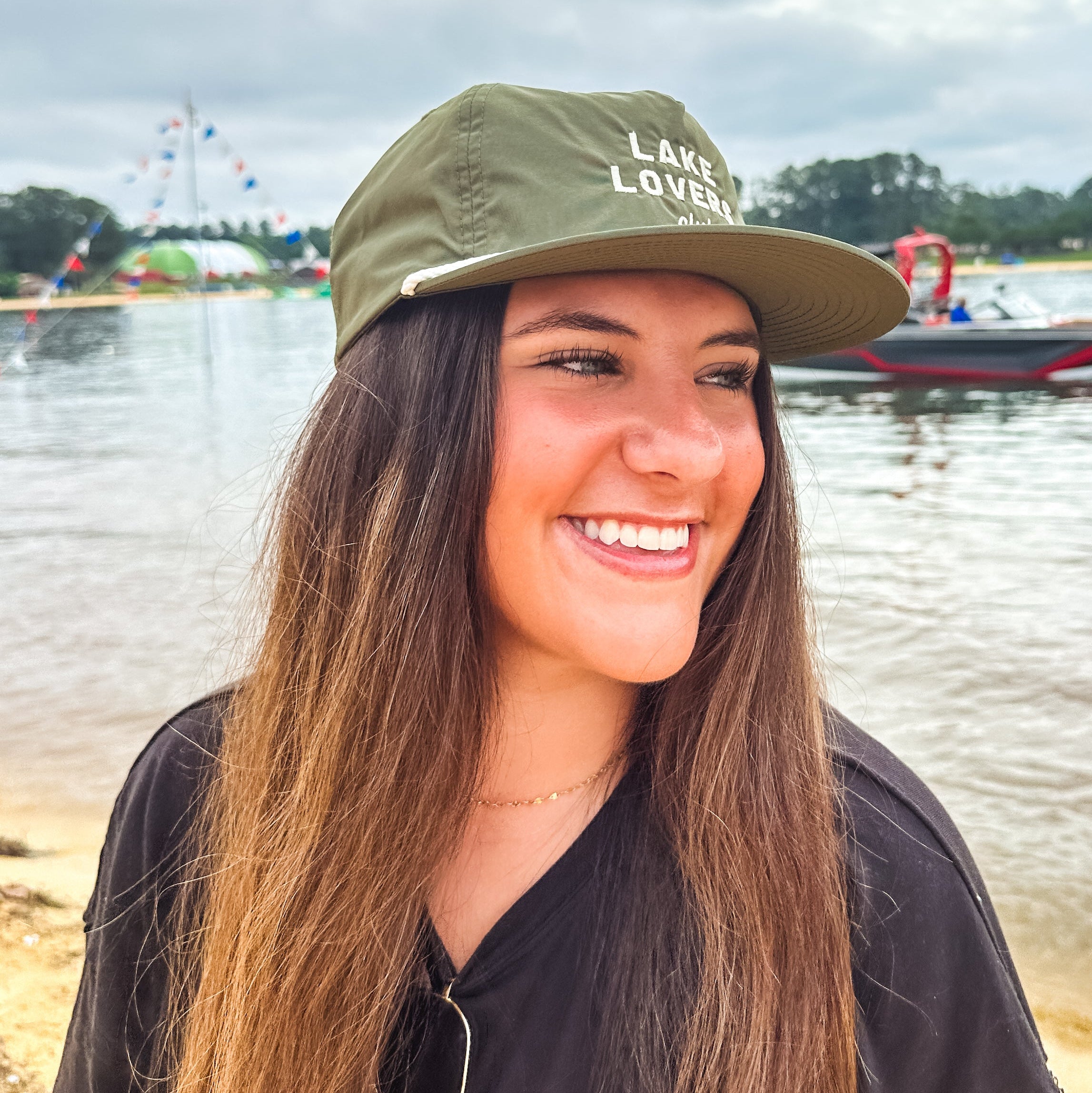 Woman wearing an olive green hat that reads 'Lake Lovers Club,' standing by a lake.