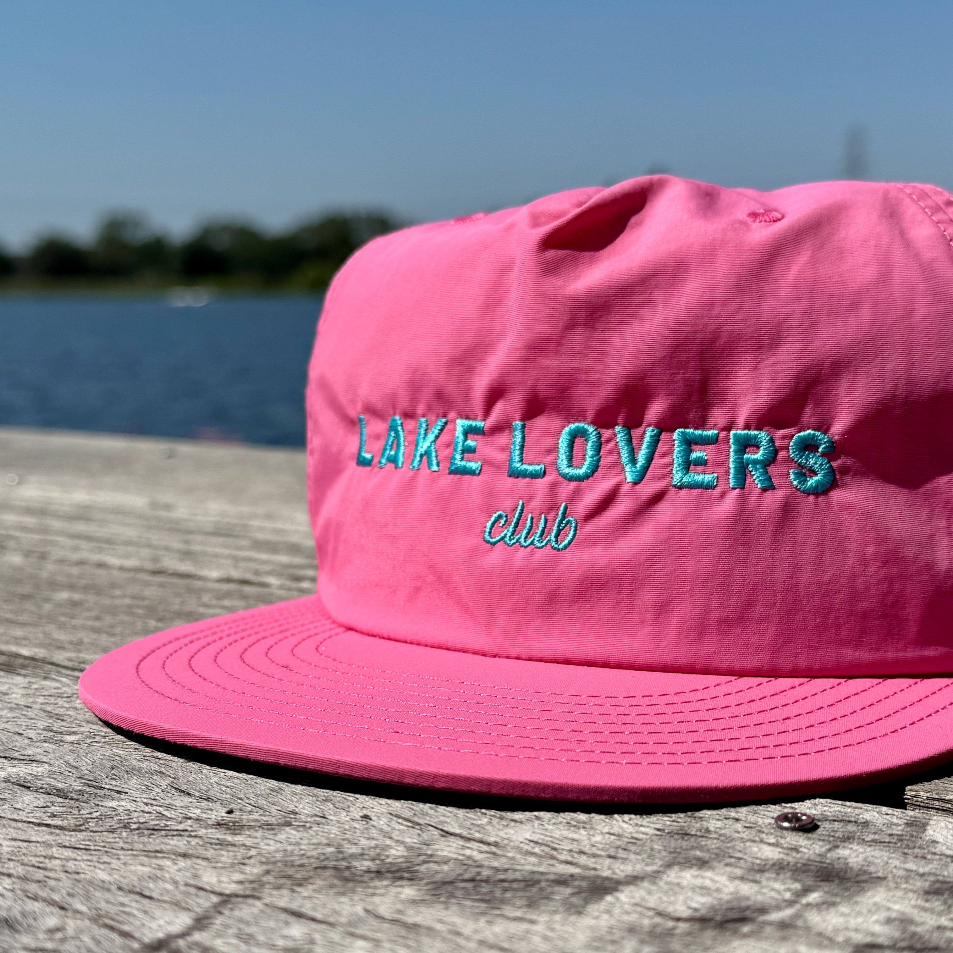 Pink cap with 'Lake Lovers Club' text on a wooden surface with a lake background