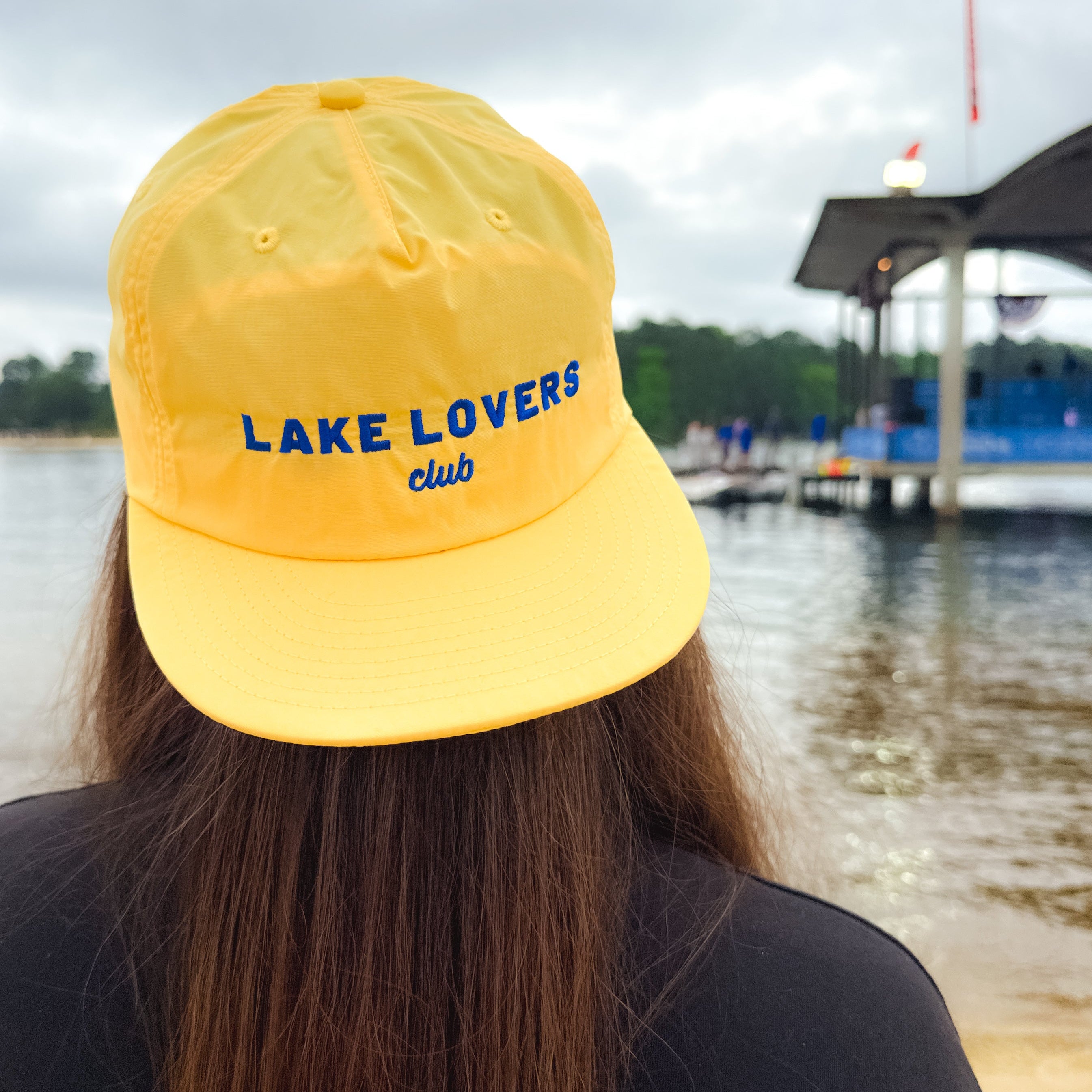 Person wearing a yellow 'Lake Lovers Club' cap by a lake with a dock in the background.