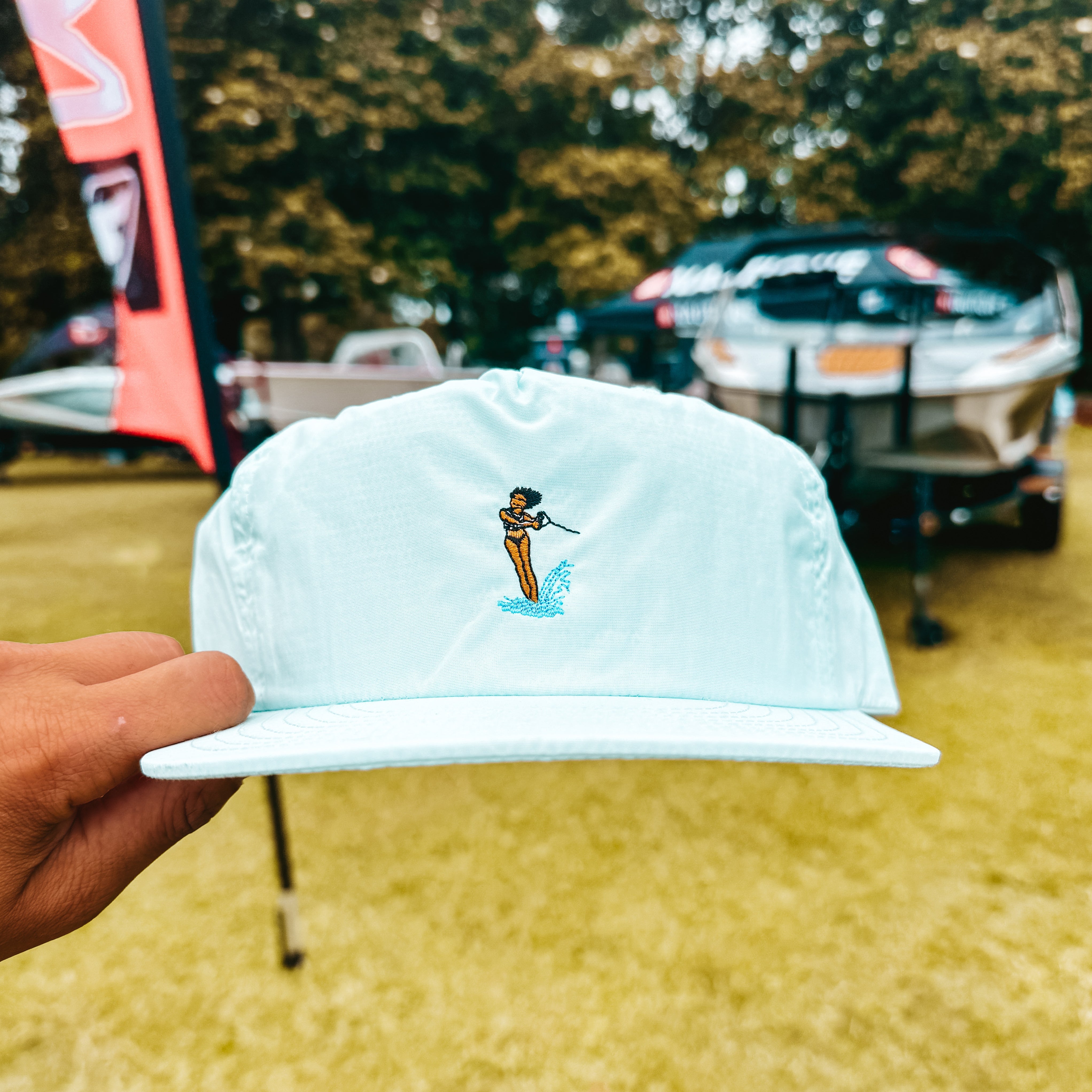 Light blue cap with a waterski design held by a hand, with blurred background of trees and wake boats.