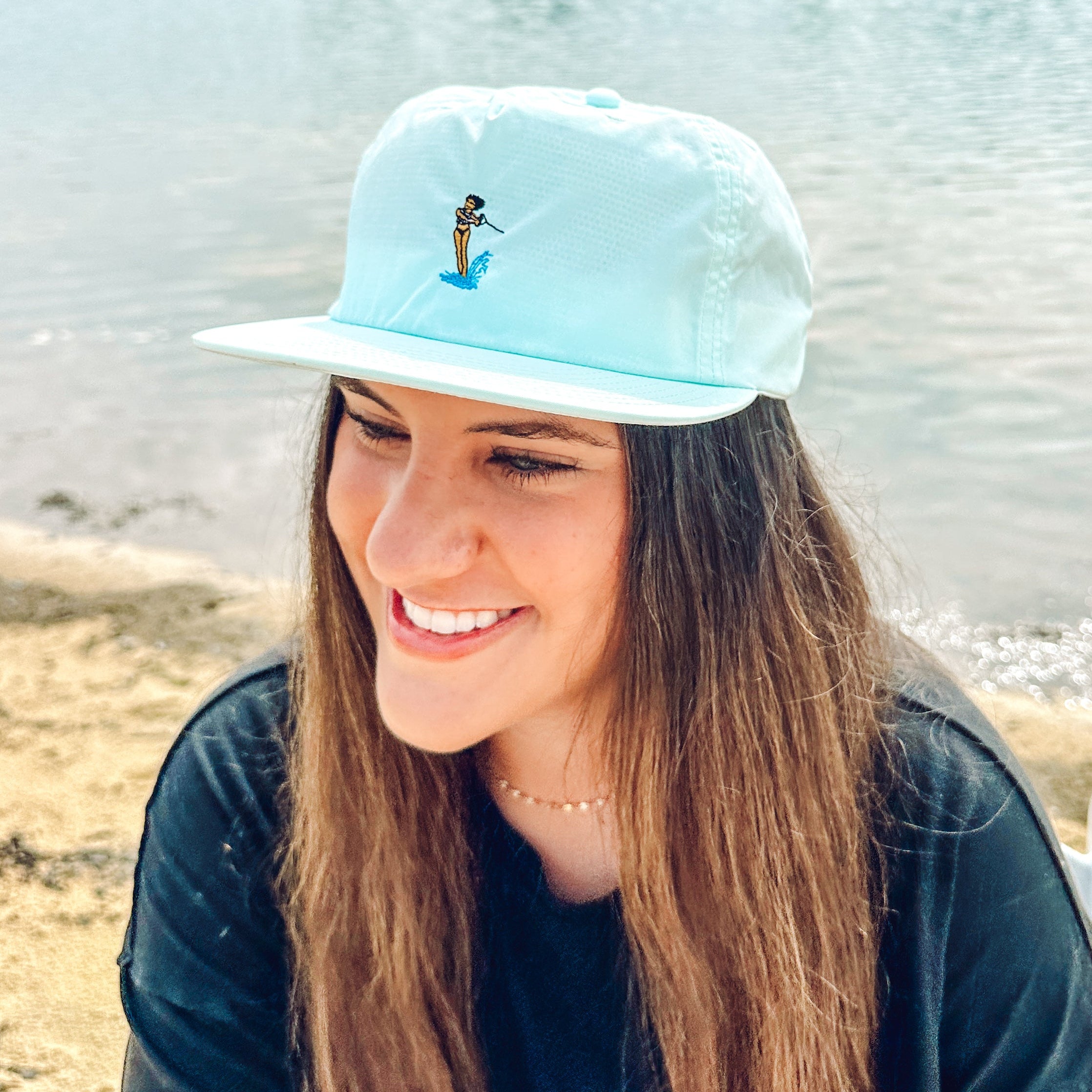 Woman wearing a light blue cap with a waterski girl design, sitting by a body of water.