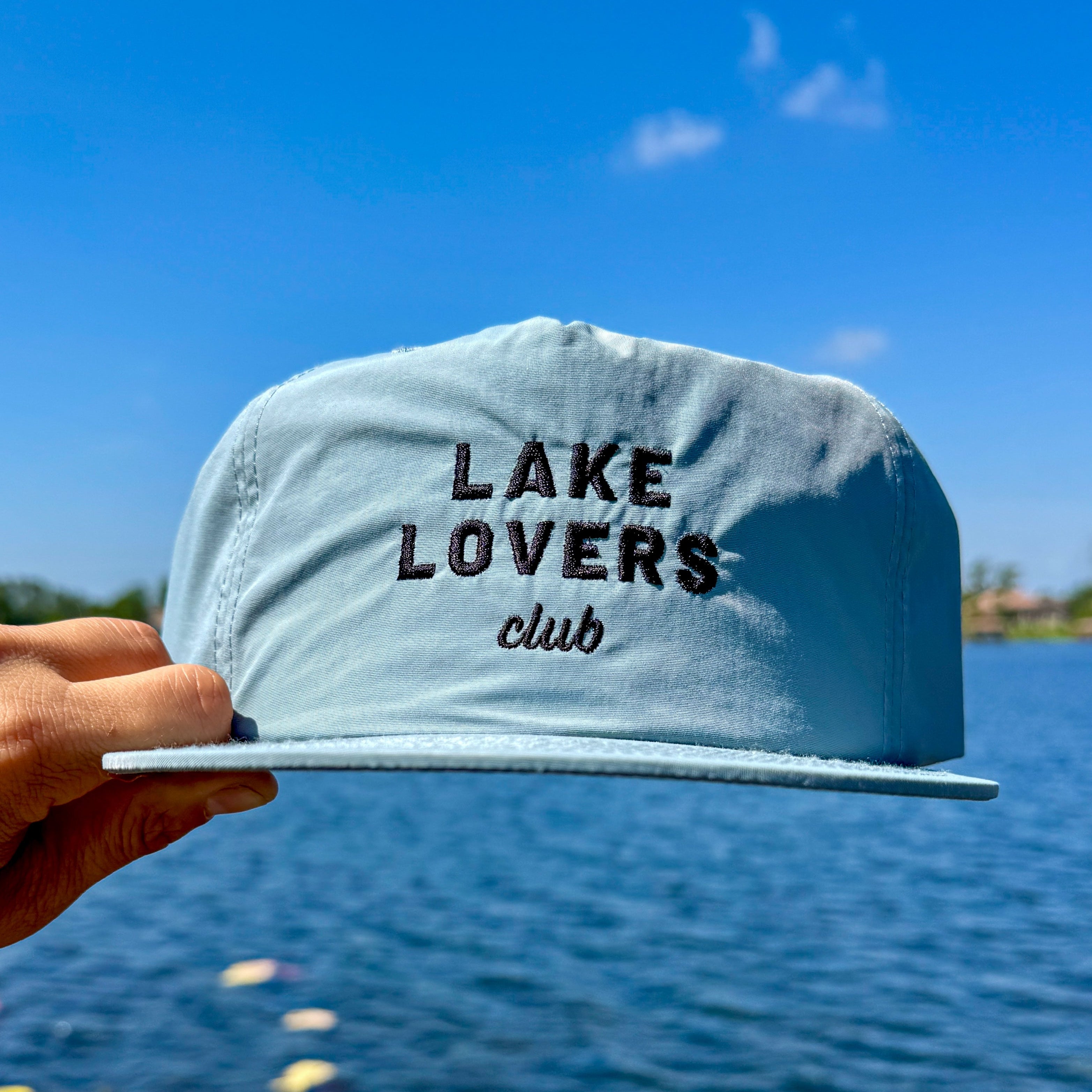 Light grey cap with 'Lake Lovers club' text held over a lake with blue sky.