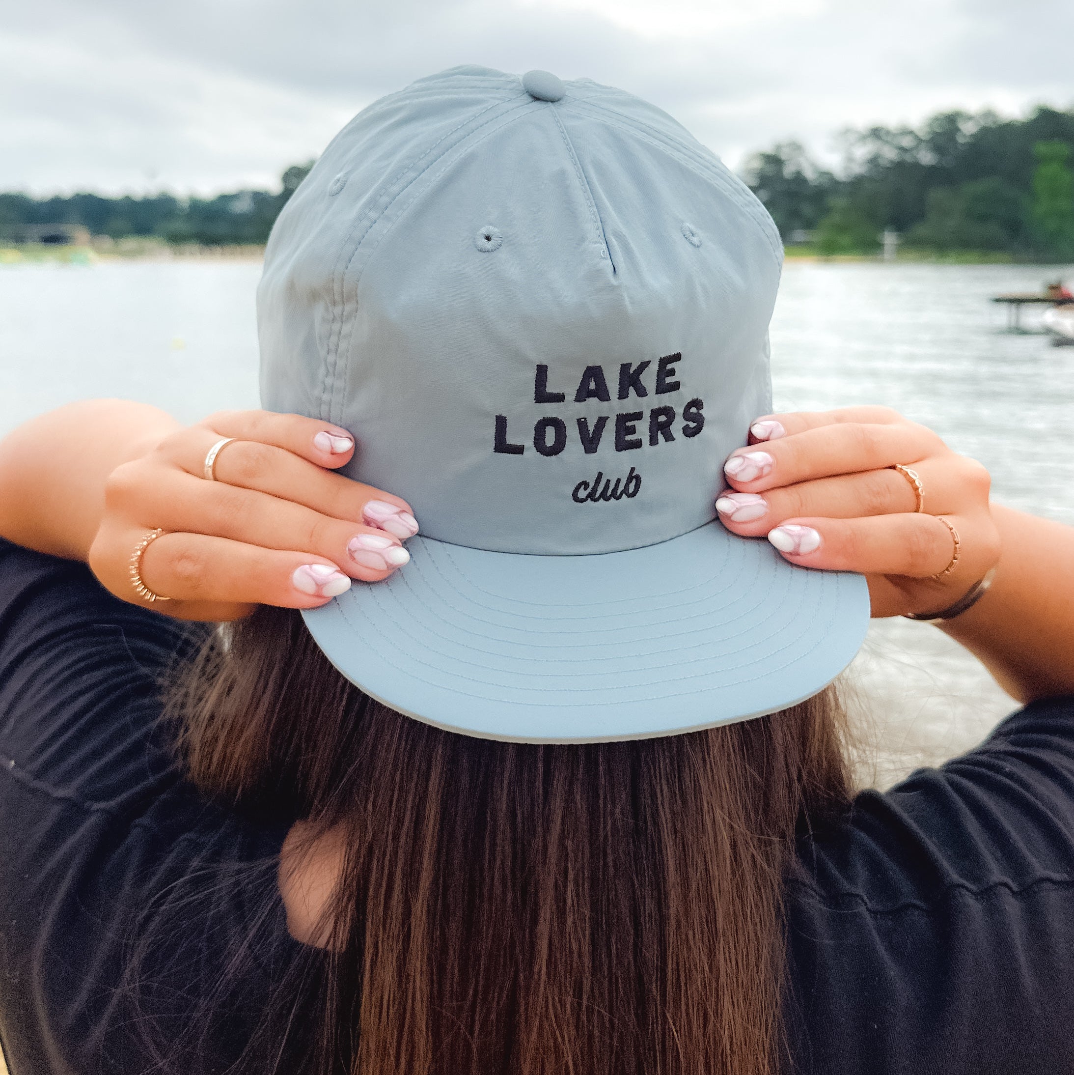 Person wearing a light grey cap with 'Lake Lovers Club' text by a lake.