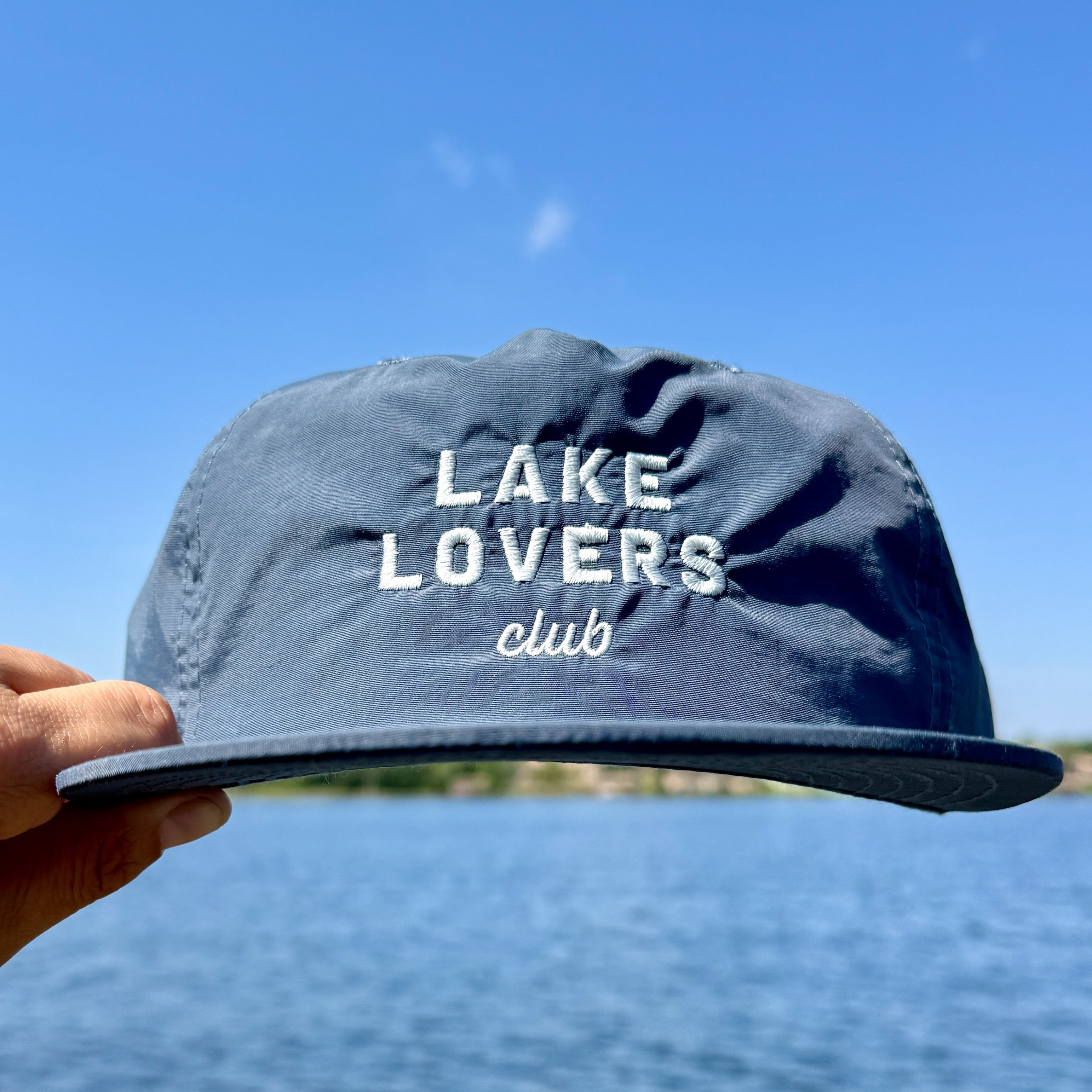 Blue cap with 'Lake Lovers Club' text held against a blue sky and water background