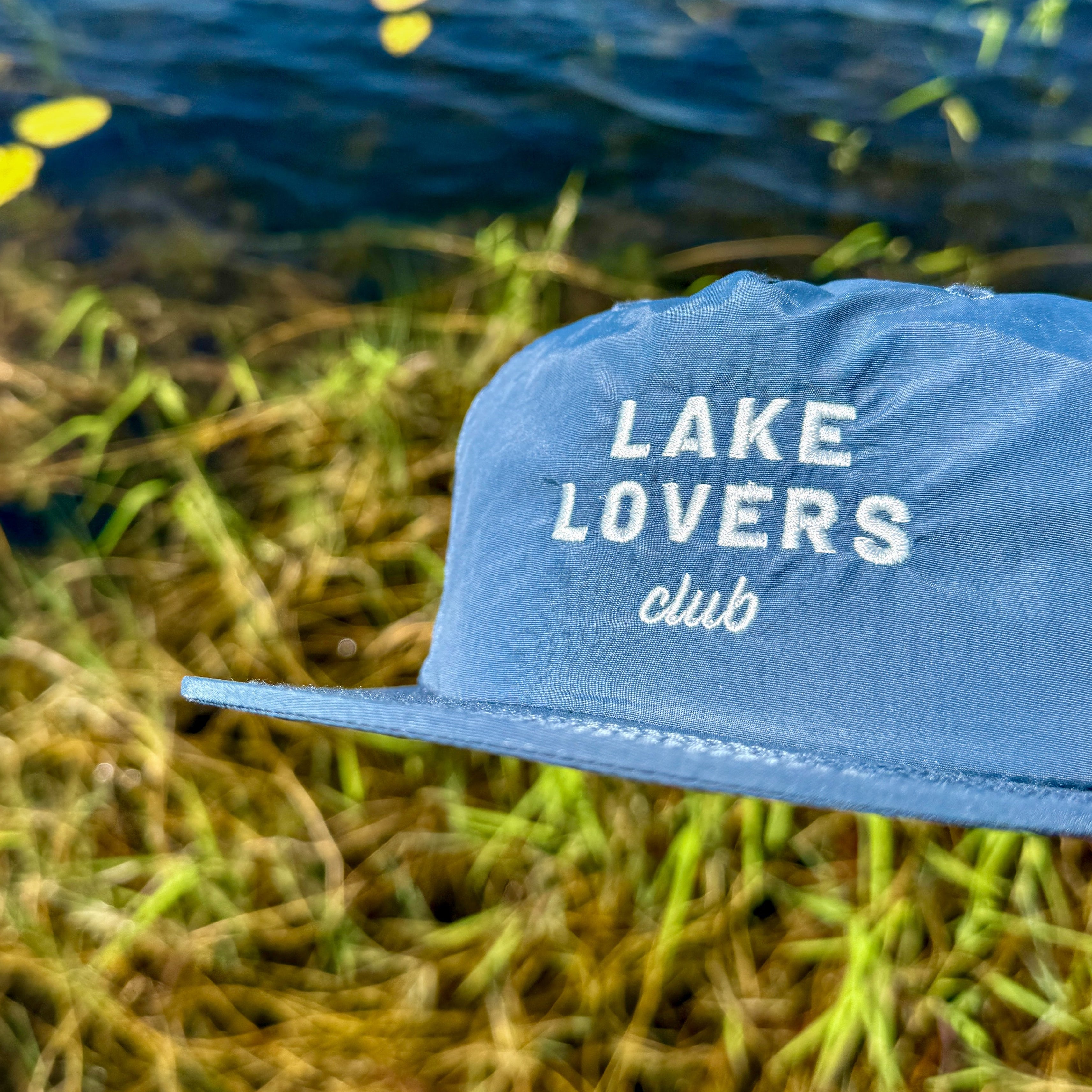 Blue cap with 'Lake Lovers club' text on grass near water
