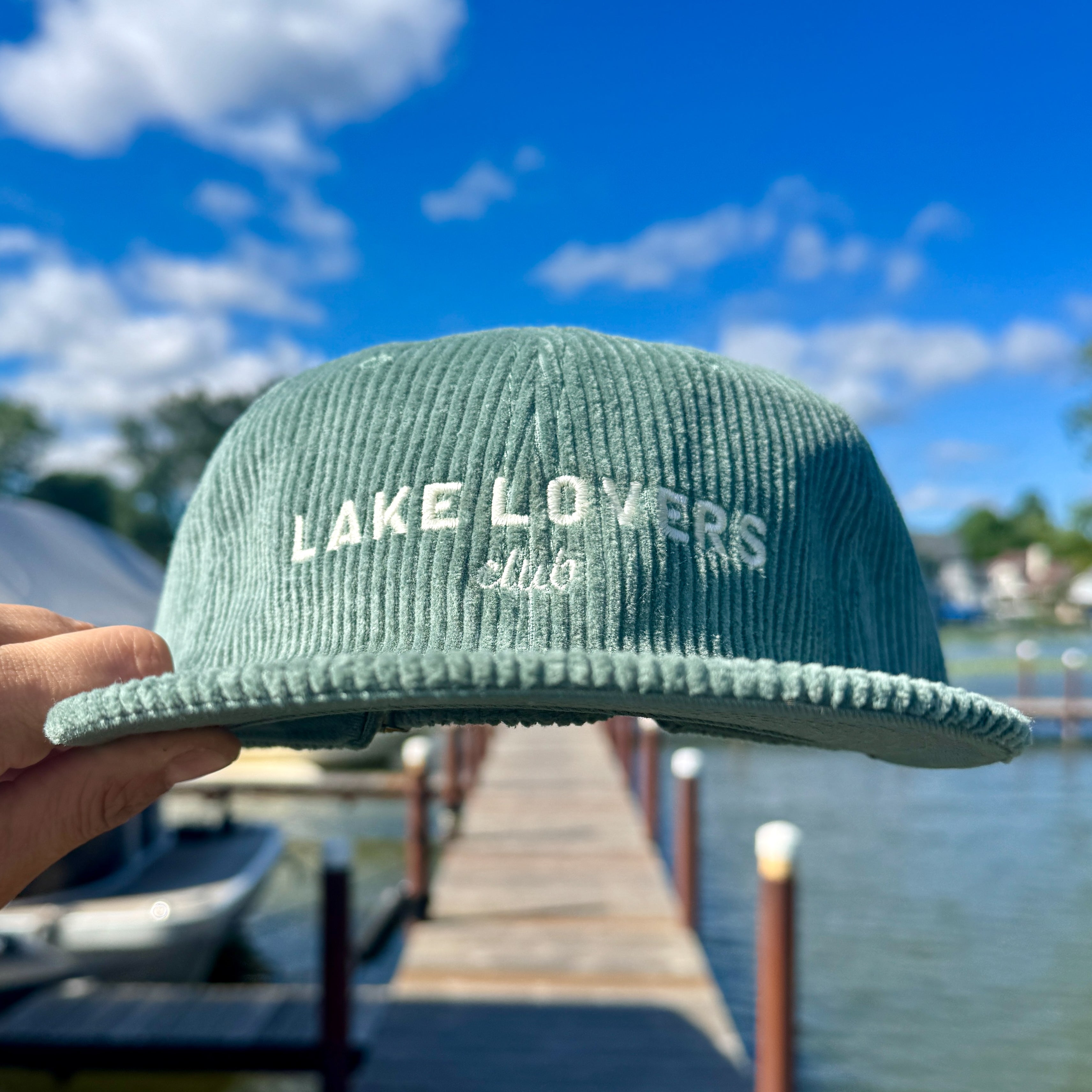 Green cap with 'Lake Lovers Club' text held by a hand with a lake and dock background