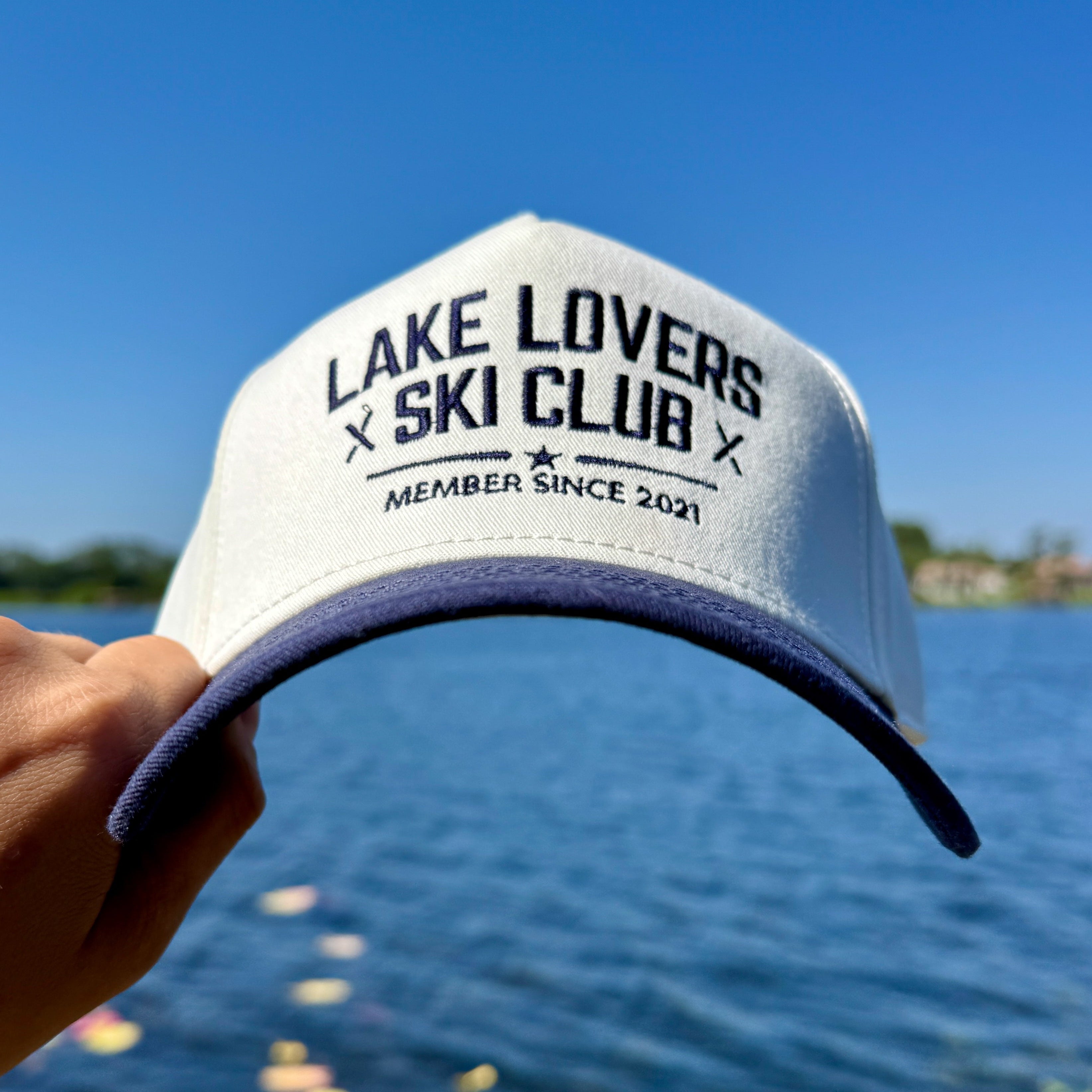 White cap with 'Lake Lovers Ski Club' text held over a lake and blue sky background