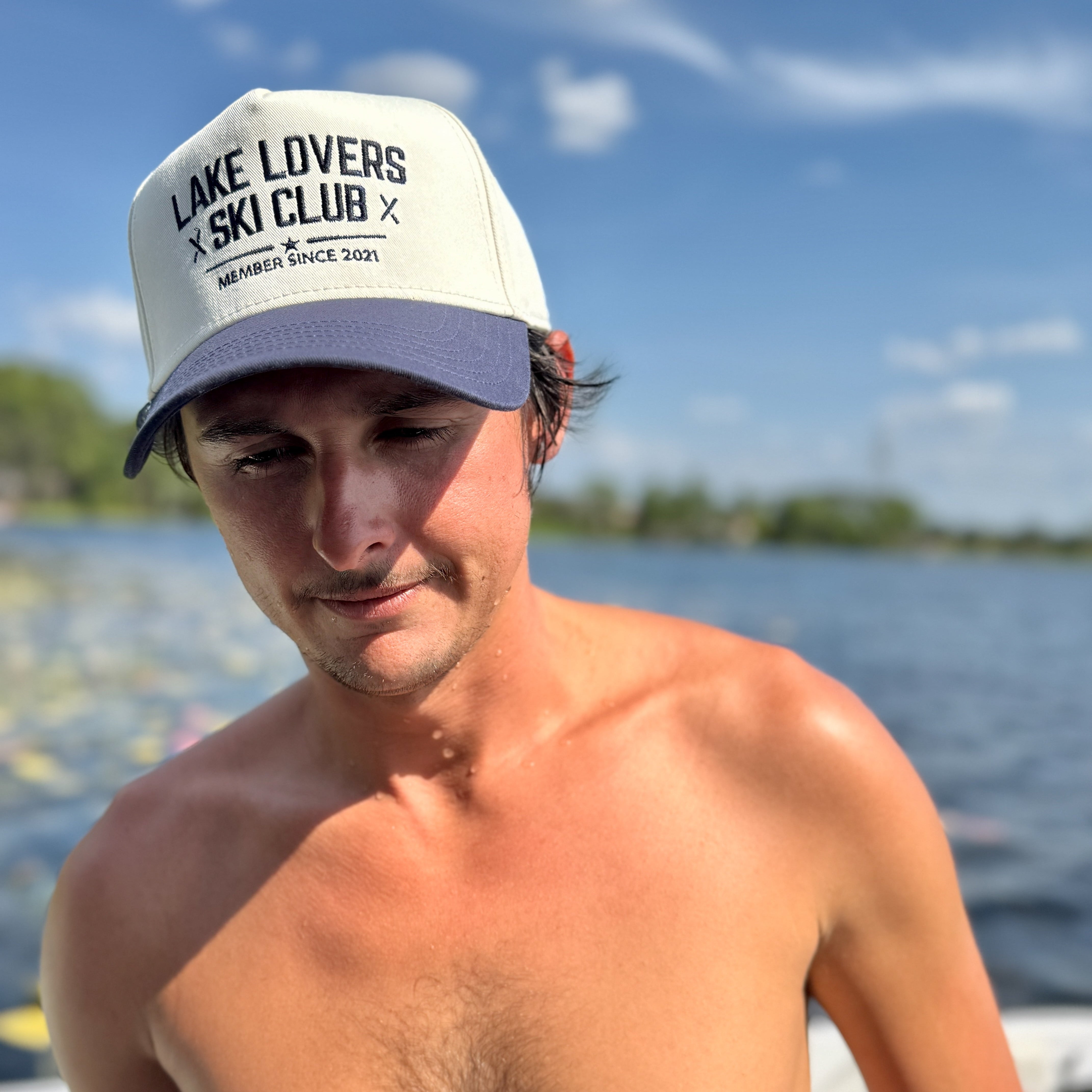 Man wearing a 'Lake Lovers Ski Club' cap by a lake