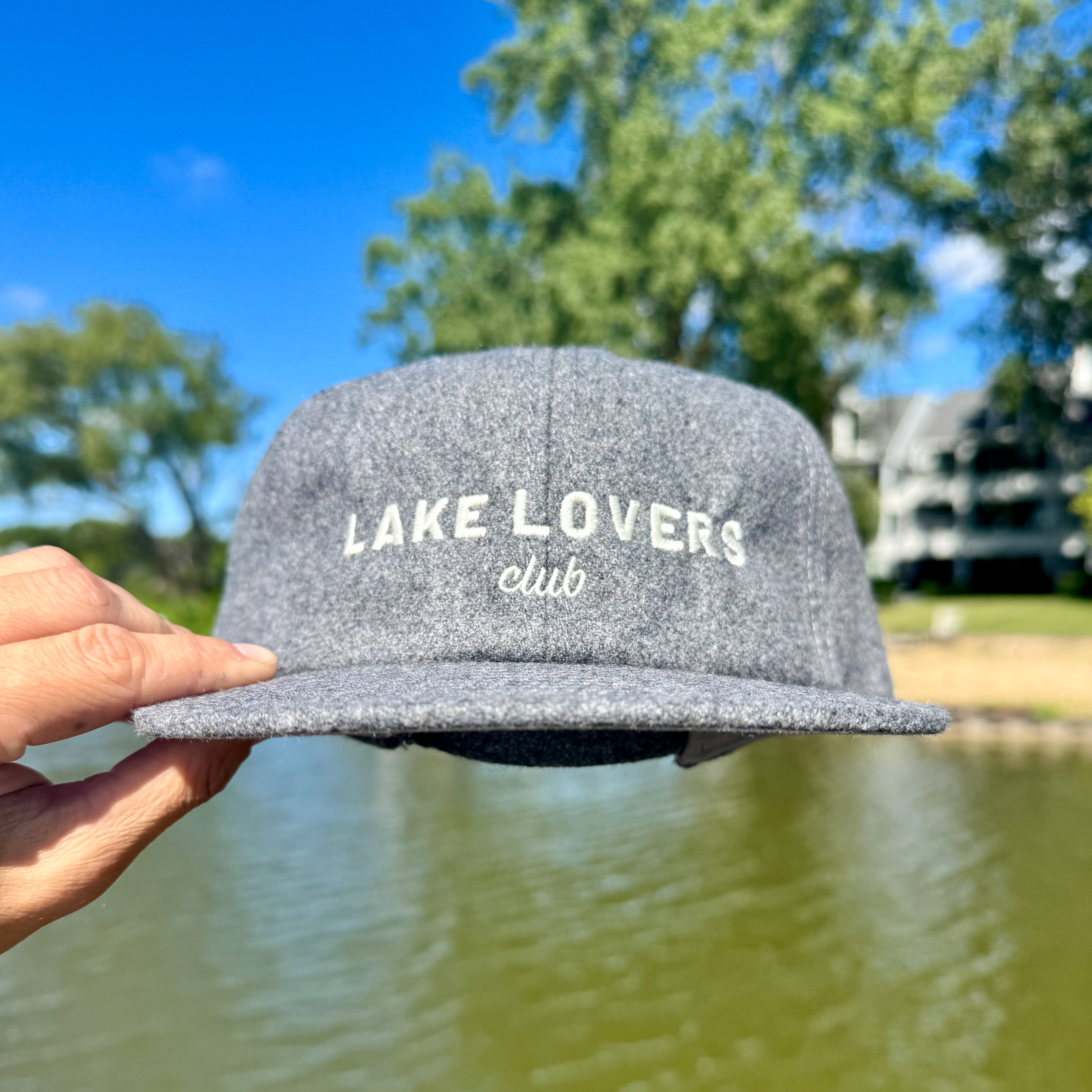Gray cap with 'Lake Lovers Club' text held by a hand against a lakeside background