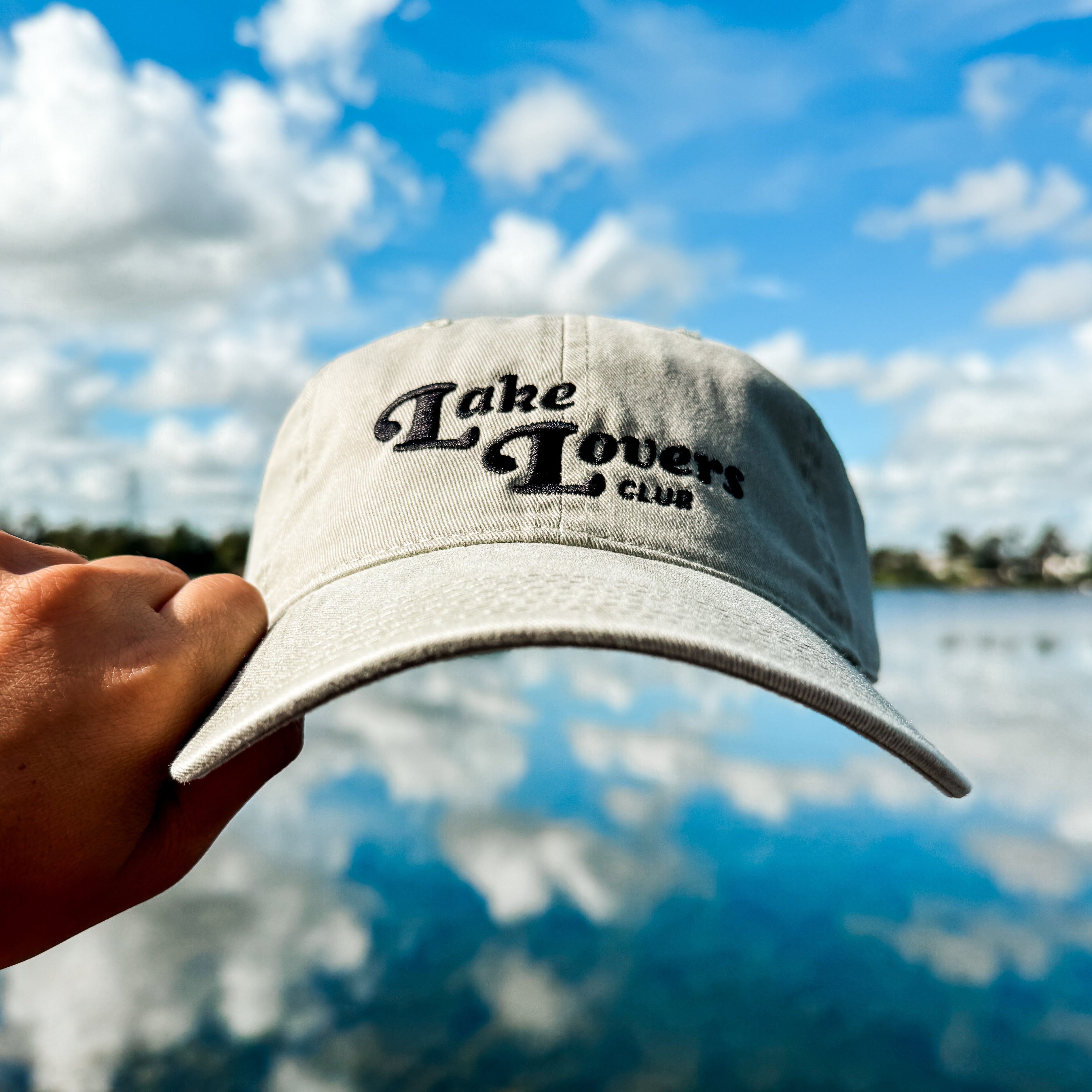 Cap with 'Lake Lover Club' text held over a lake with a blue sky.