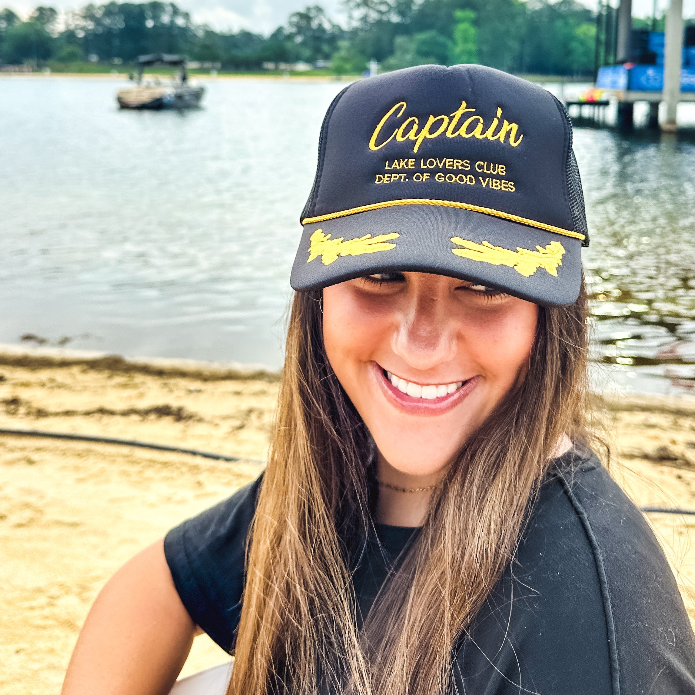 Woman wearing a 'Captain' cap by a lake with a boat in the background