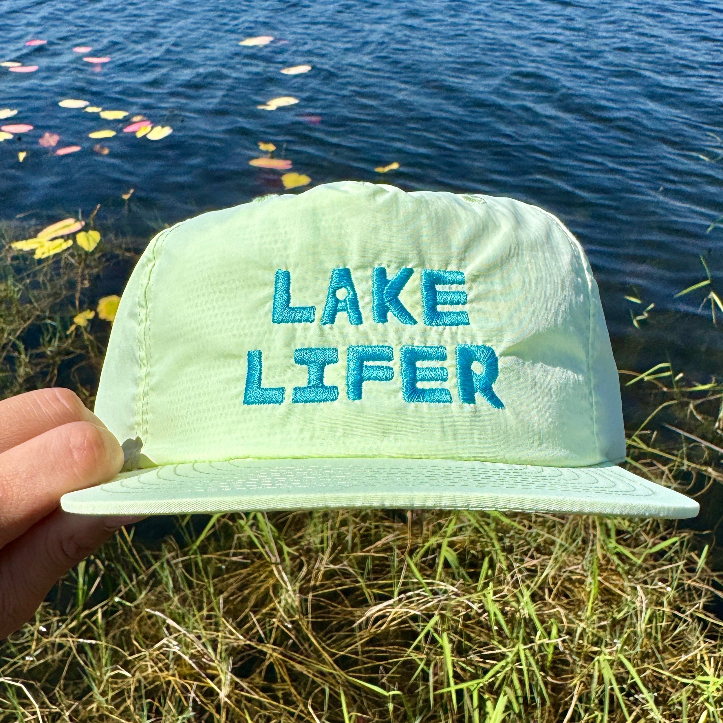 Light green cap with 'Lake Lifer' text held over a lake with water and grass in the background
