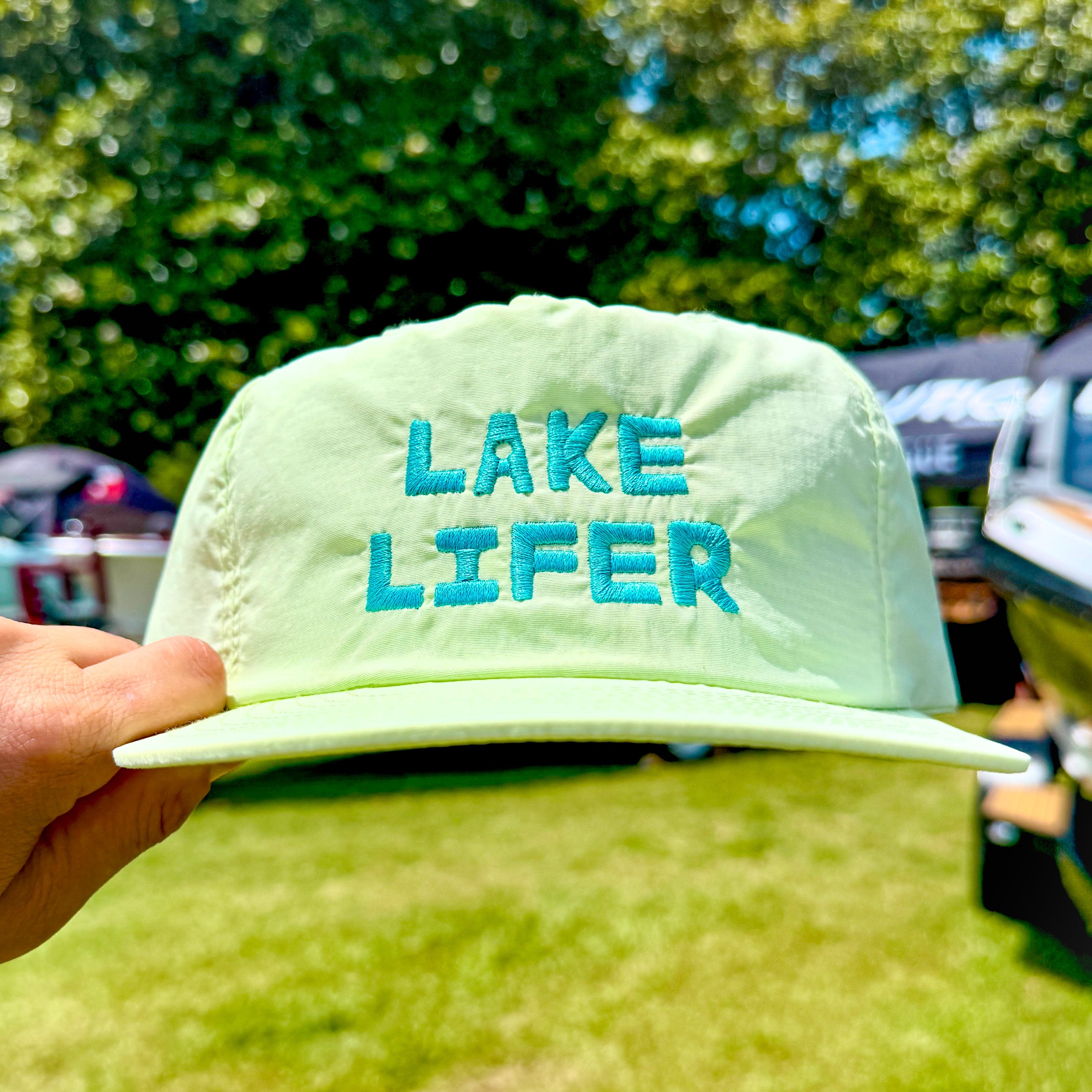 Light green cap with 'Lake Lifer' text held in front of a blurred outdoor background