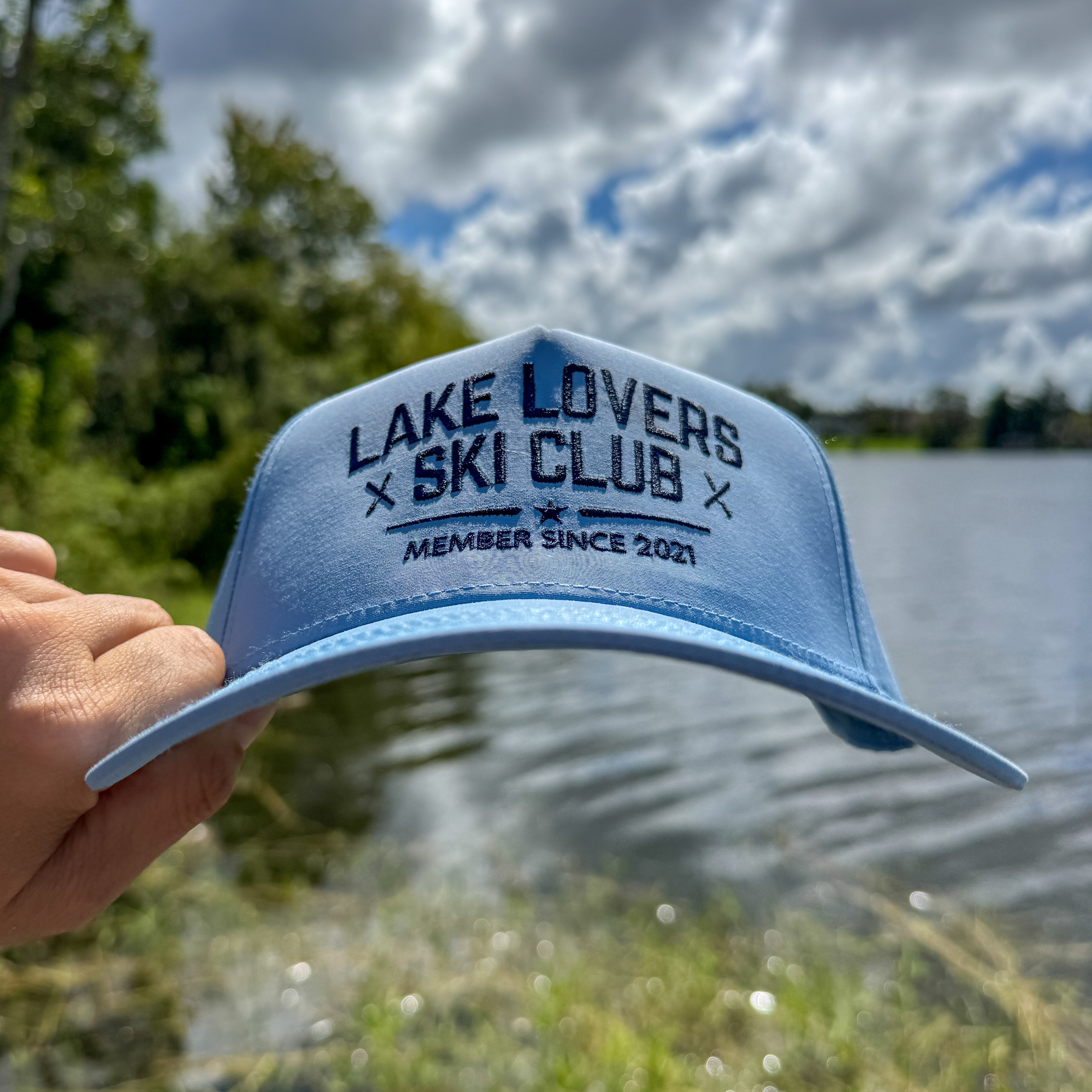 Blue cap with 'Lake Lovers Ski Club' text held over a lake and trees