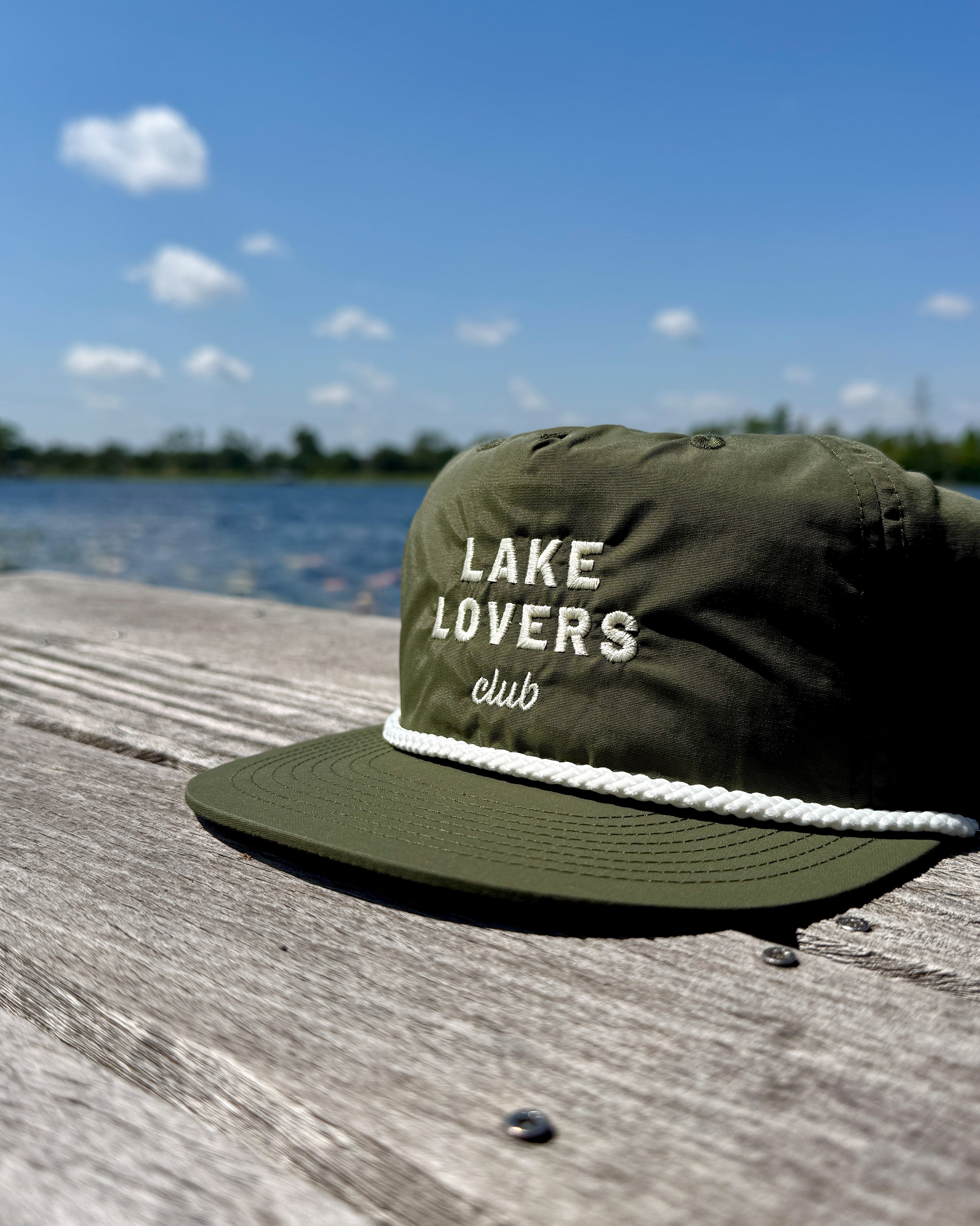 Green cap with 'Lake Lovers Club' text on a wooden dock with a lake and blue sky in the background