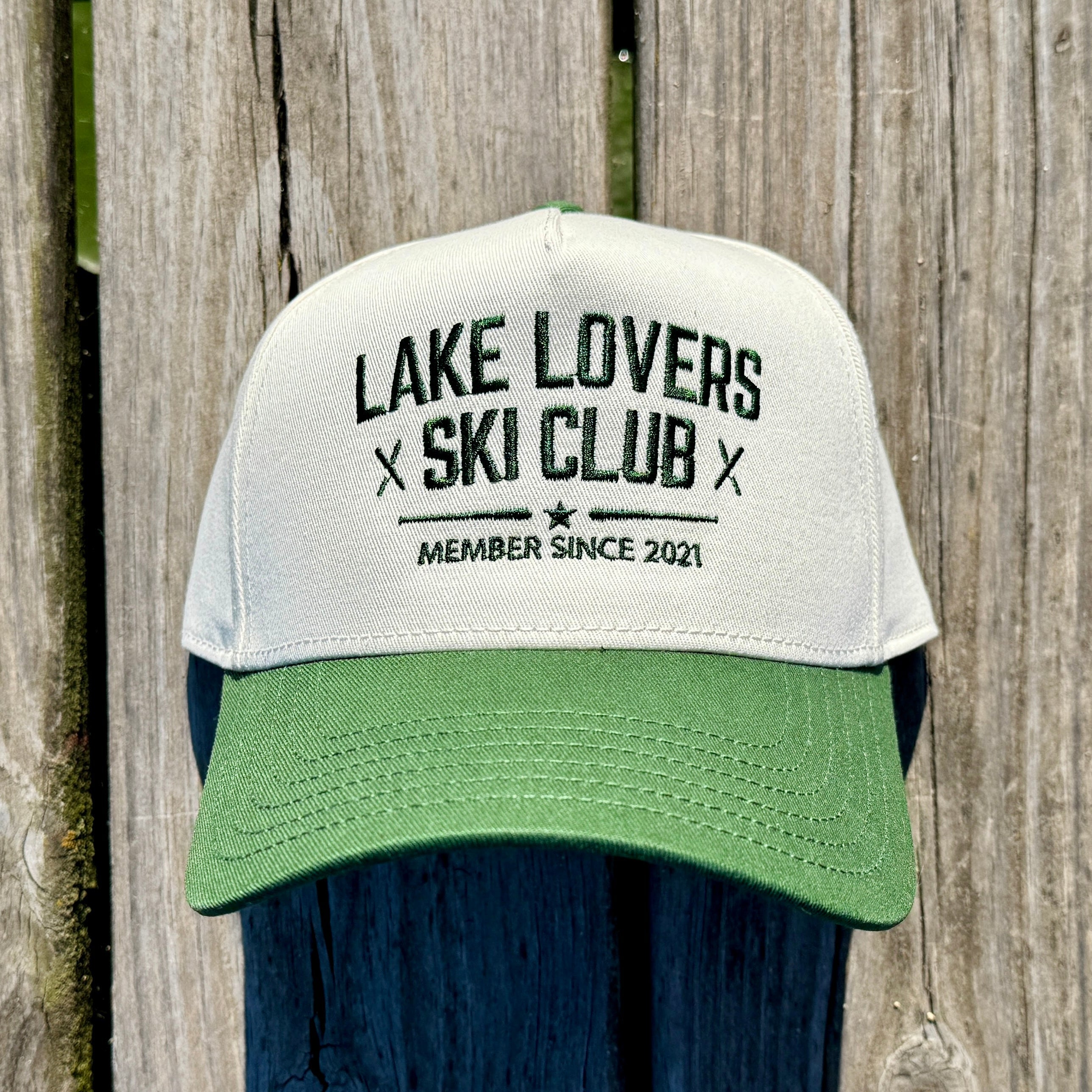 Beige hat on a dock with a forest green brim and text on it in green that says Lake Lovers Ski Club.