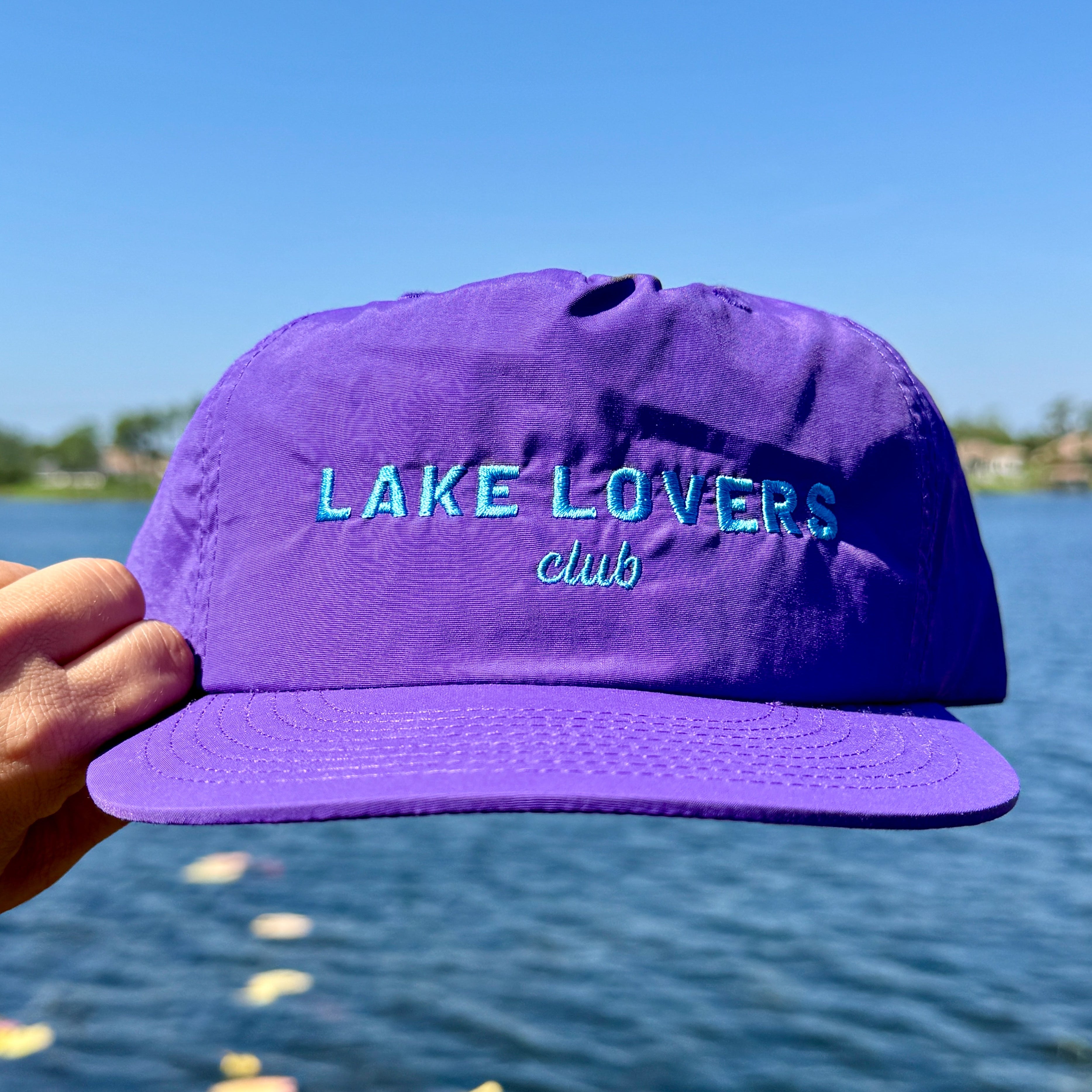 Purple cap with 'Lake Lovers Club' text held in front of a lake