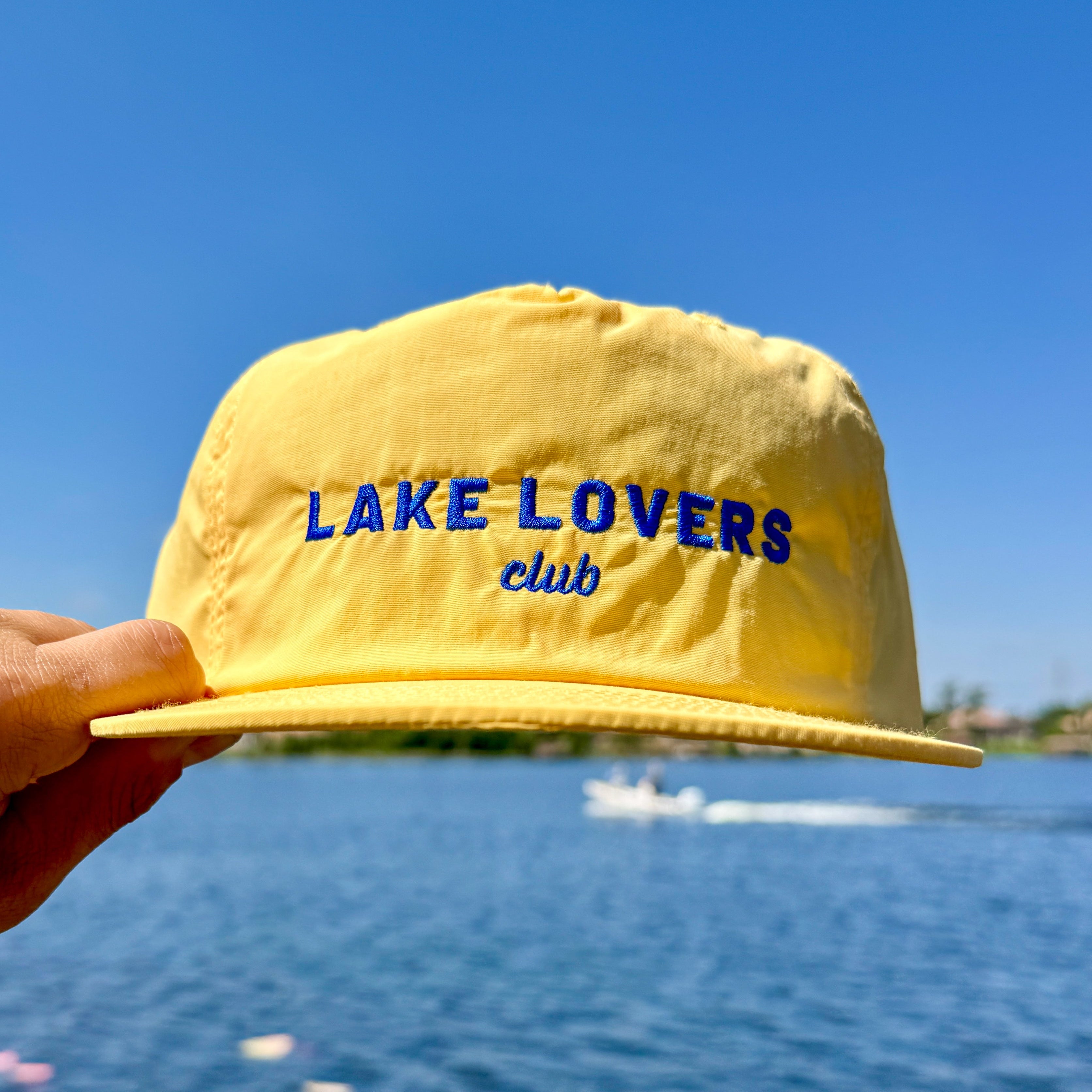 Yellow cap with 'Lake Lovers Club' text held in front of a lake and blue sky.