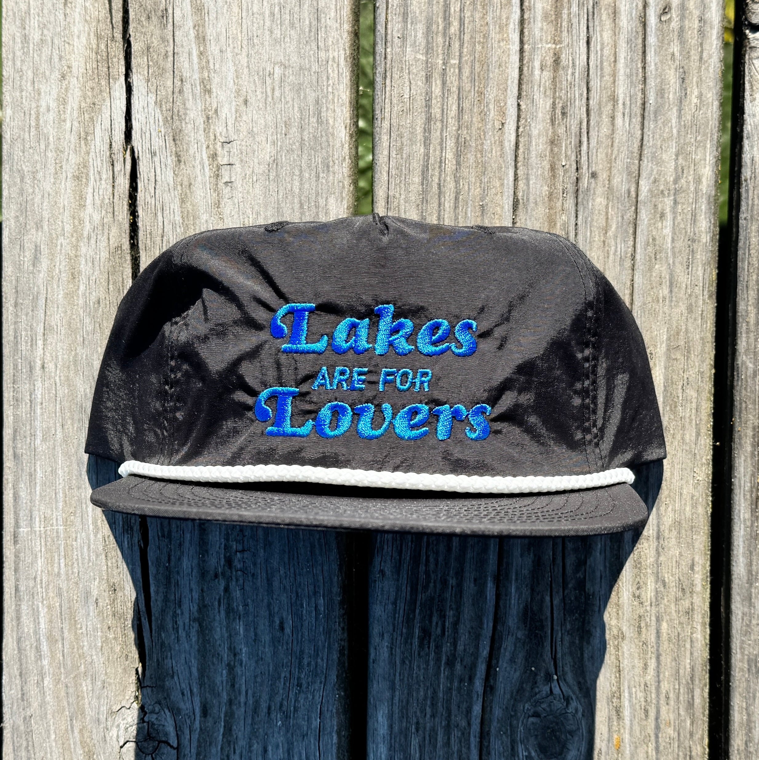 A black quick dry nylon rope hat sitting on a dock. That hat says Lakes are for Lovers in blue thread.