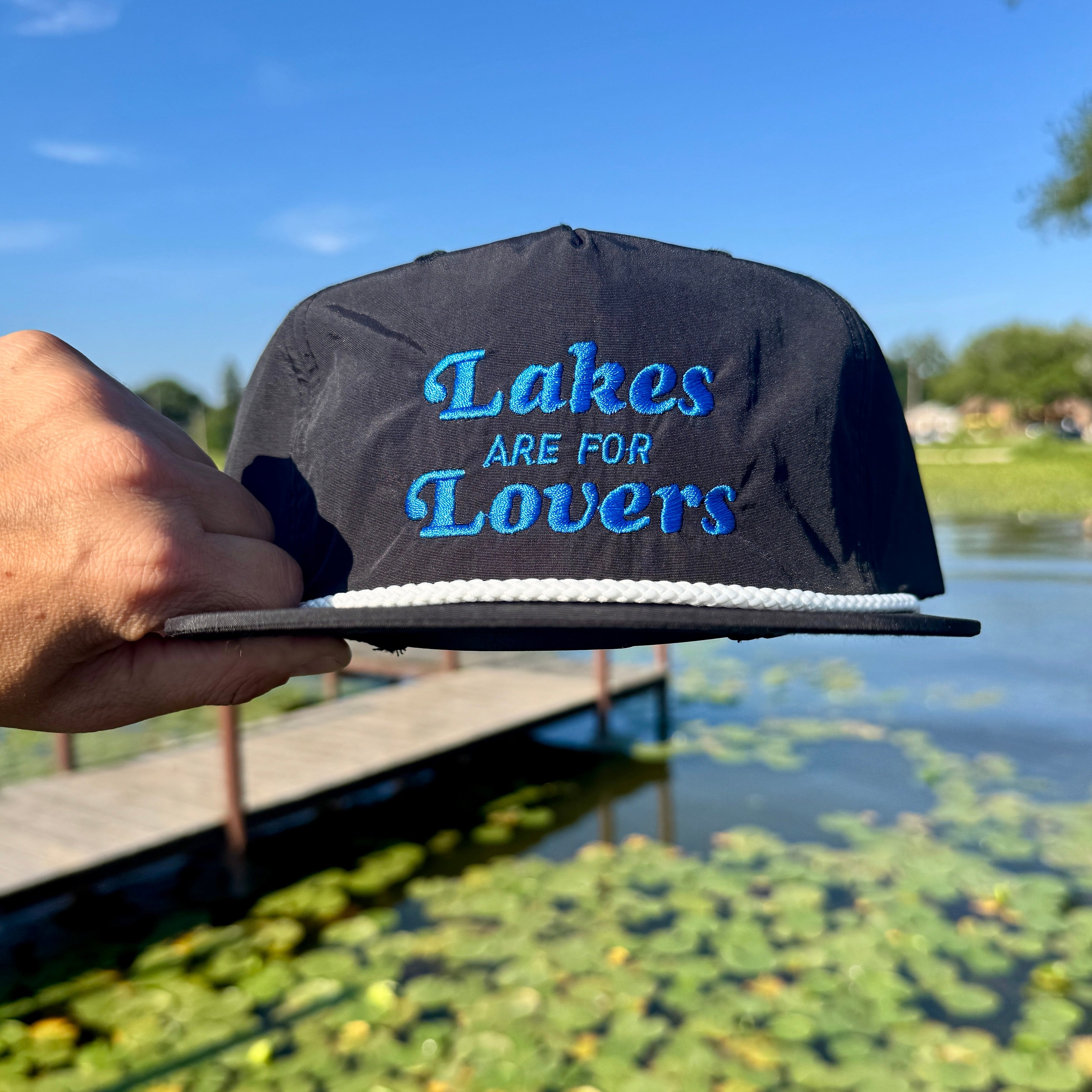 Hand holding a black quick dry nylon rope hat over water that says Lakes are for Lovers in blue thread.