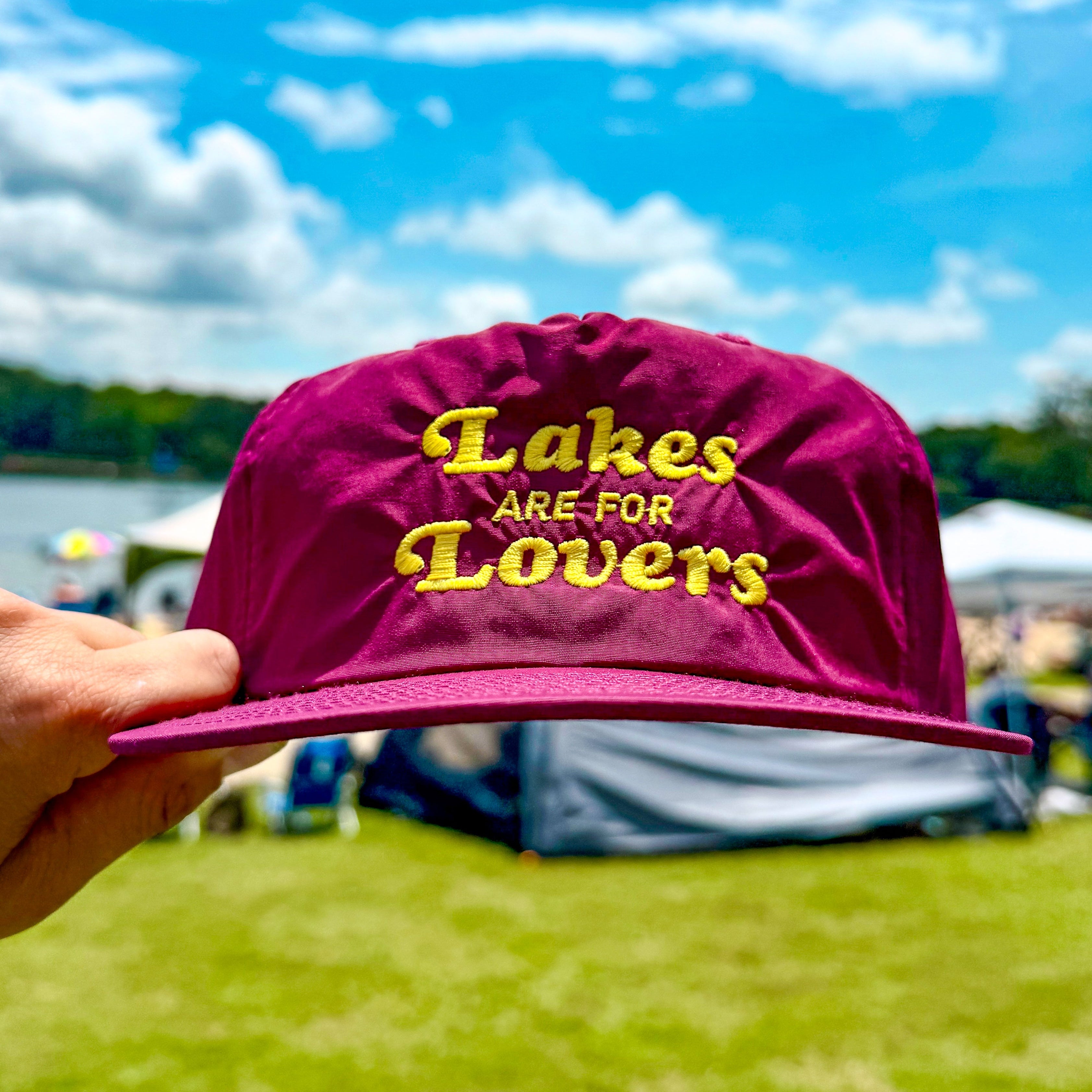Maroon cap with 'Lakes Are For Lovers' text held in front of a lakeside scene.