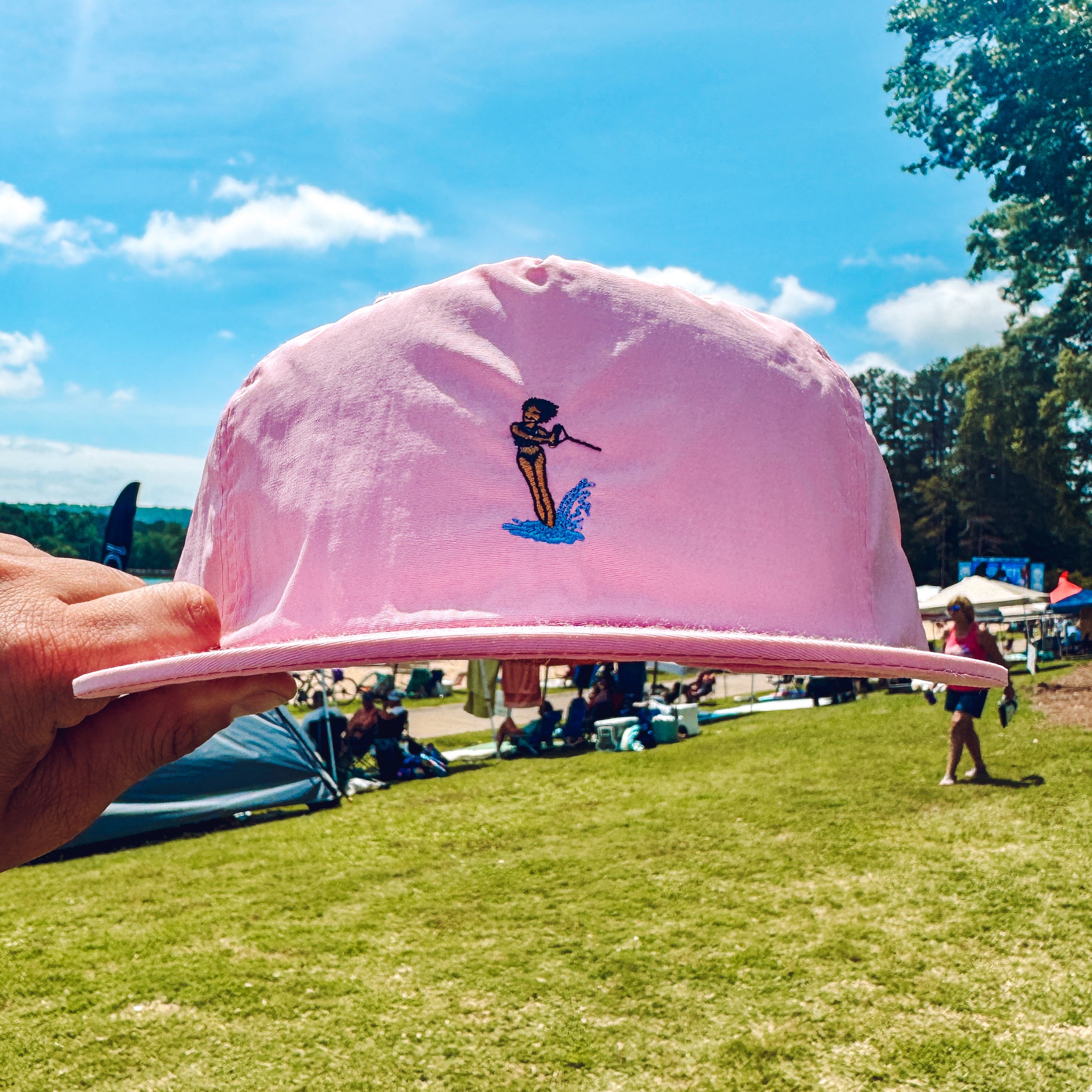 Pink cap with a waterski girl design held up against a blue sky and grassy field background
