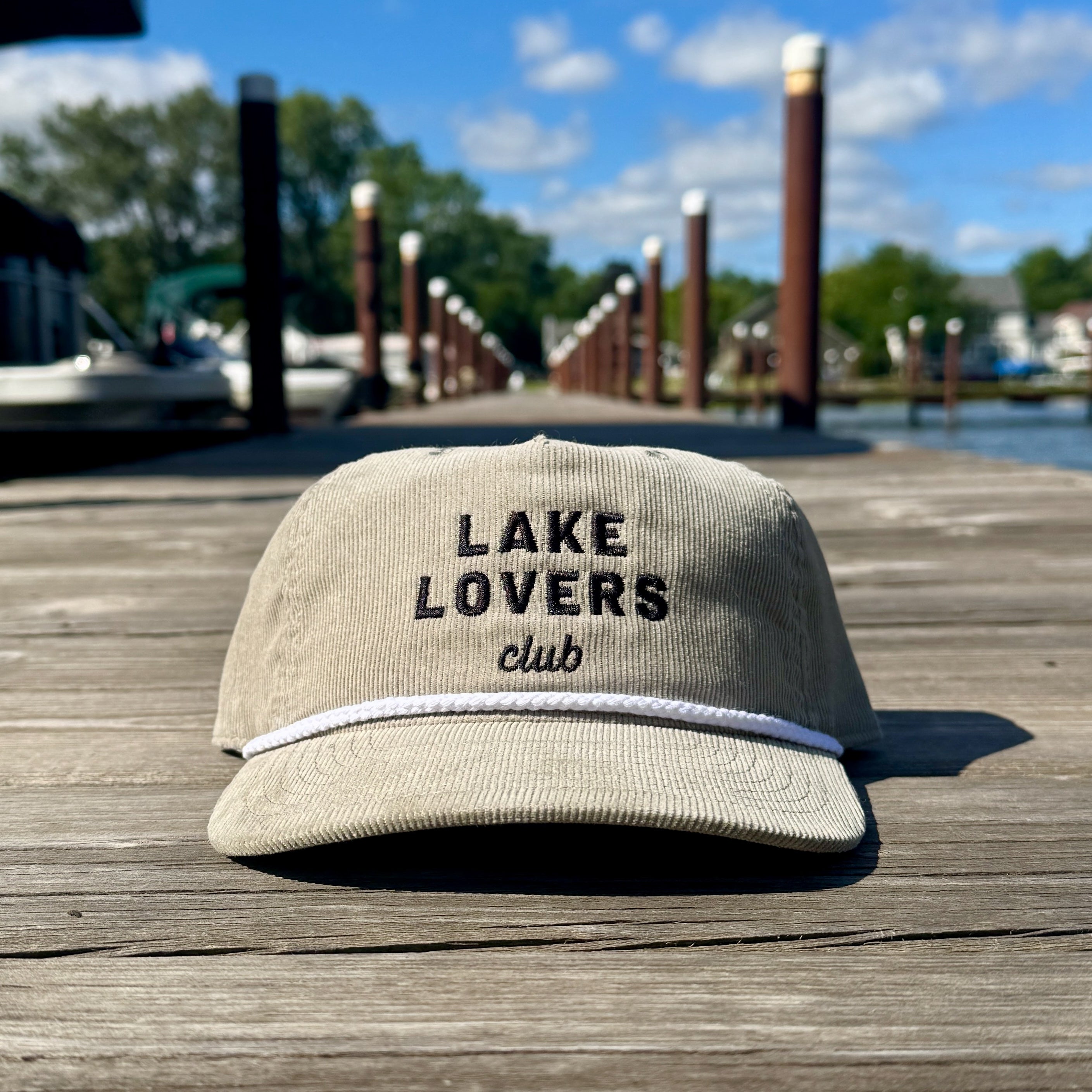 Beige cap with 'Lake Lovers club' text on a wooden dock with water and docks in the background