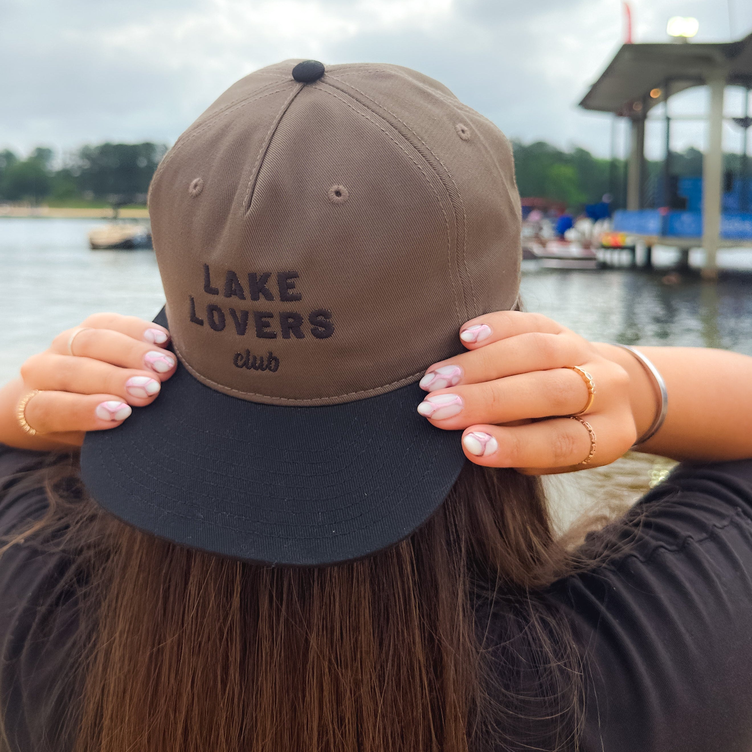Person wearing a cap with 'Lake Lovers Club' text by a lake